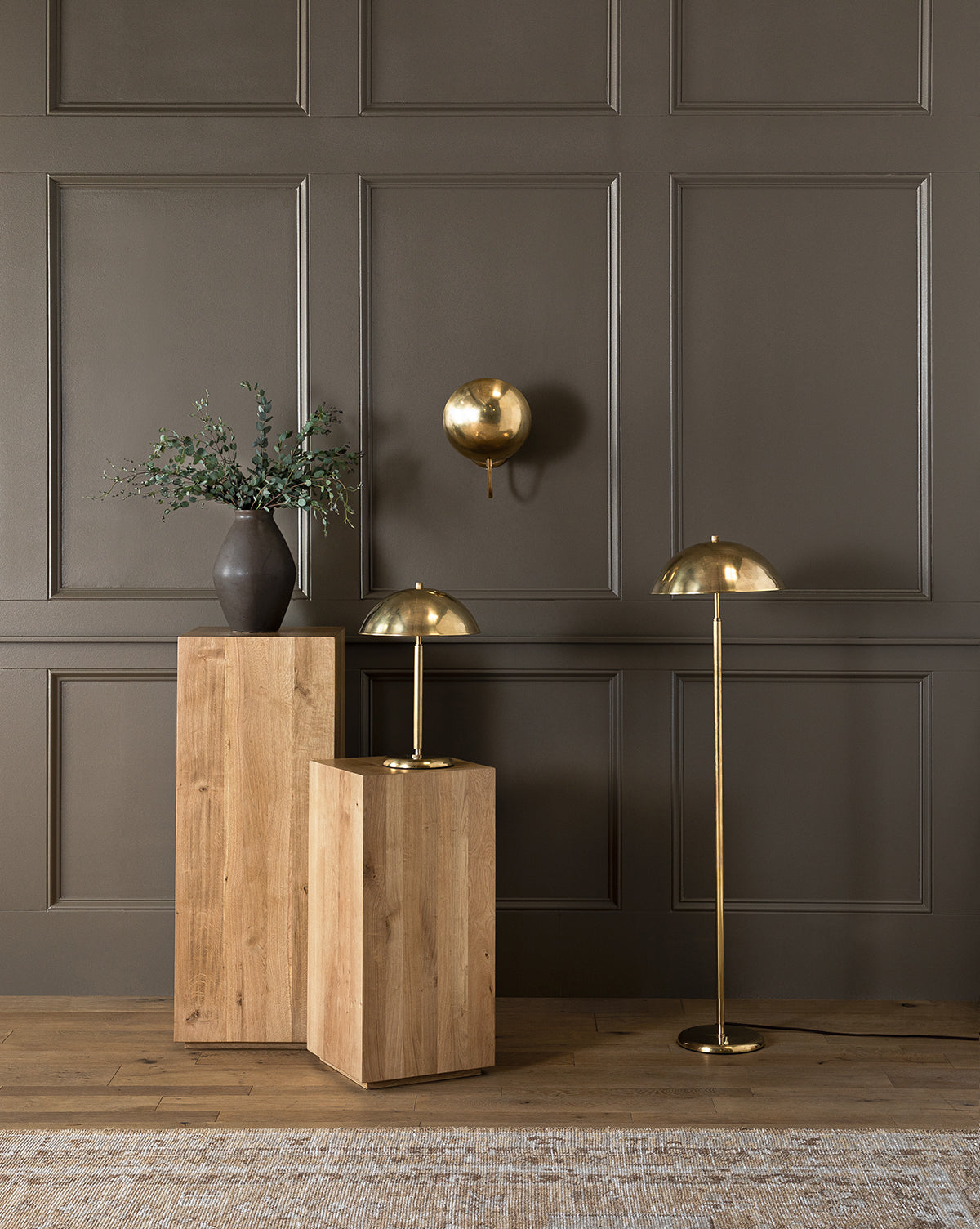 A modern interior with a dark paneled wall, two wooden pedestals, a black vase with greenery, and three gold dome-shaped lamps—including the elegant Aldorno Floor Lamp—creates a sophisticated, stylish atmosphere, by McGee & Co.