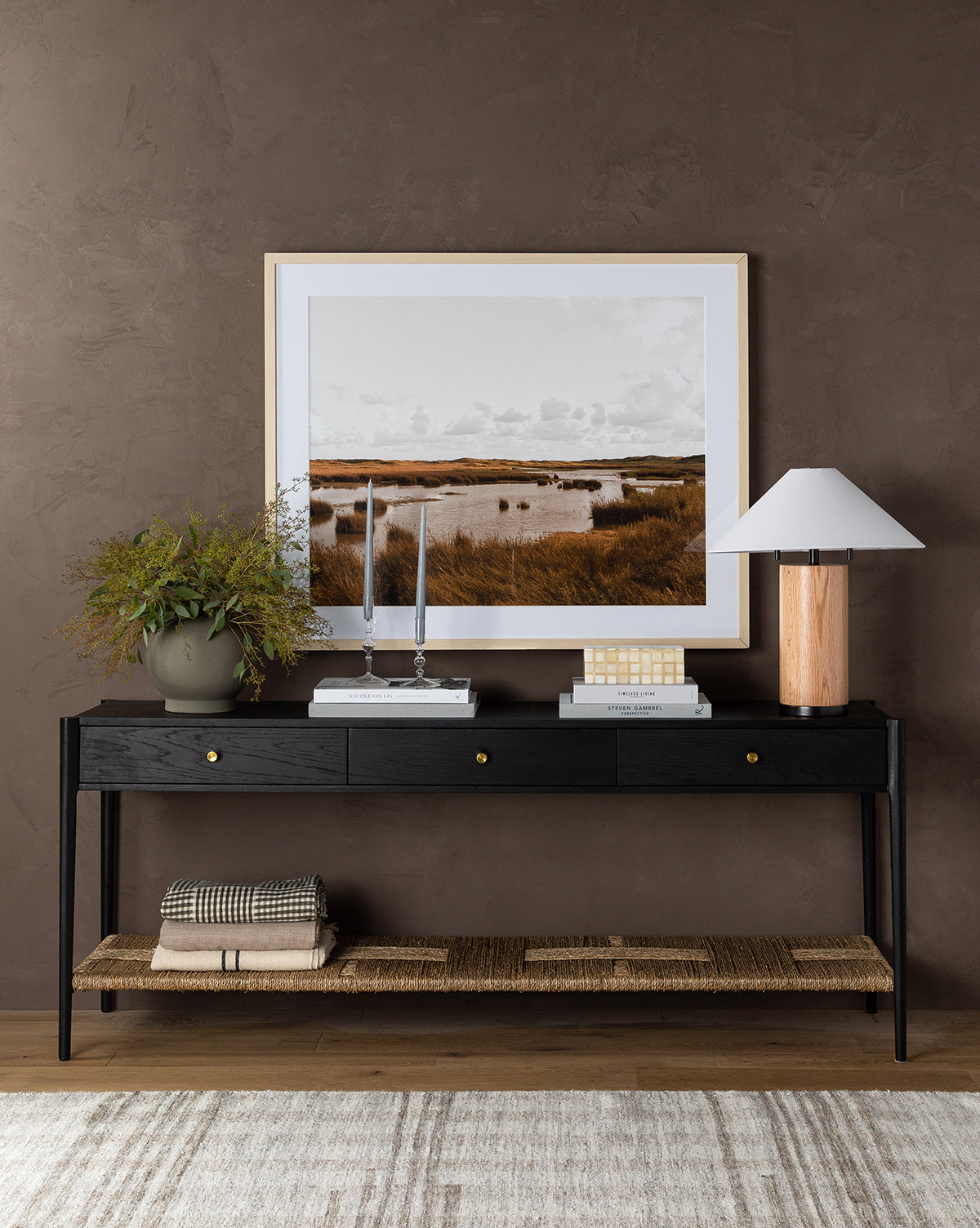 The Dana Console in black with brass hardware holds a lamp, vase with plant, candlesticks, books, and decor. A framed landscape photo hangs above. Blankets are folded on the woven shelf below—a perfect entryway accent, by McGee & Co.
