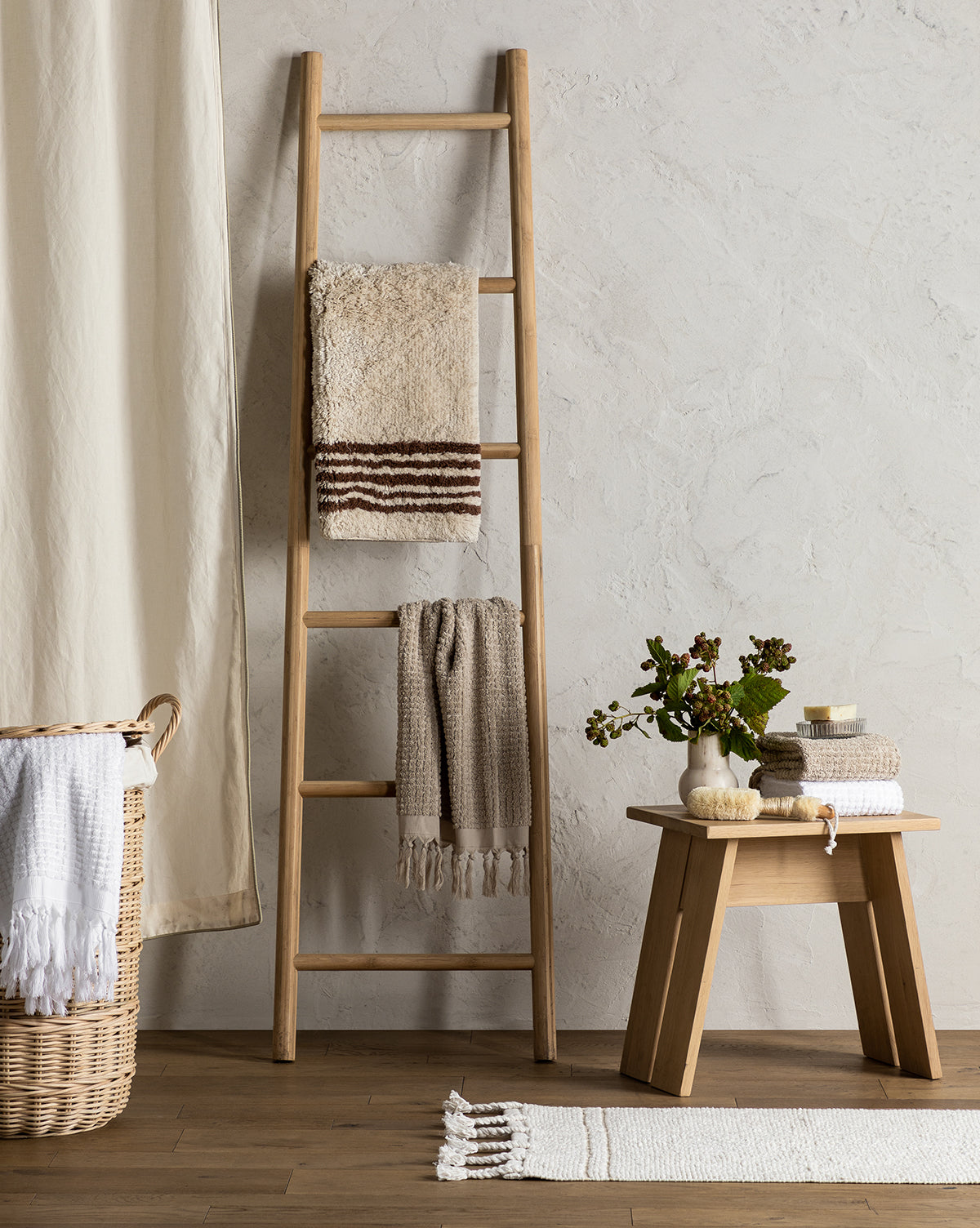 A wooden ladder with neutral-toned towels leans against a textured wall. A wicker basket with a white towel, the Beata Bath Mat, and a wooden bench topped with folded towels, soap, and greenery complete the cozy minimalist scene.