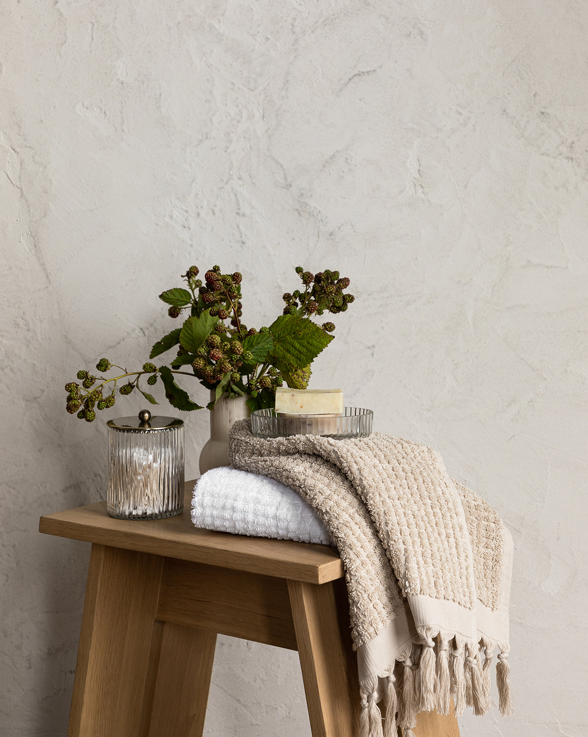 A wooden stool displays folded beige and white towels, a jar from the Stein Glass Collection, a soap dish with soap, and a small vase with green leaves and berries against a textured light wall.