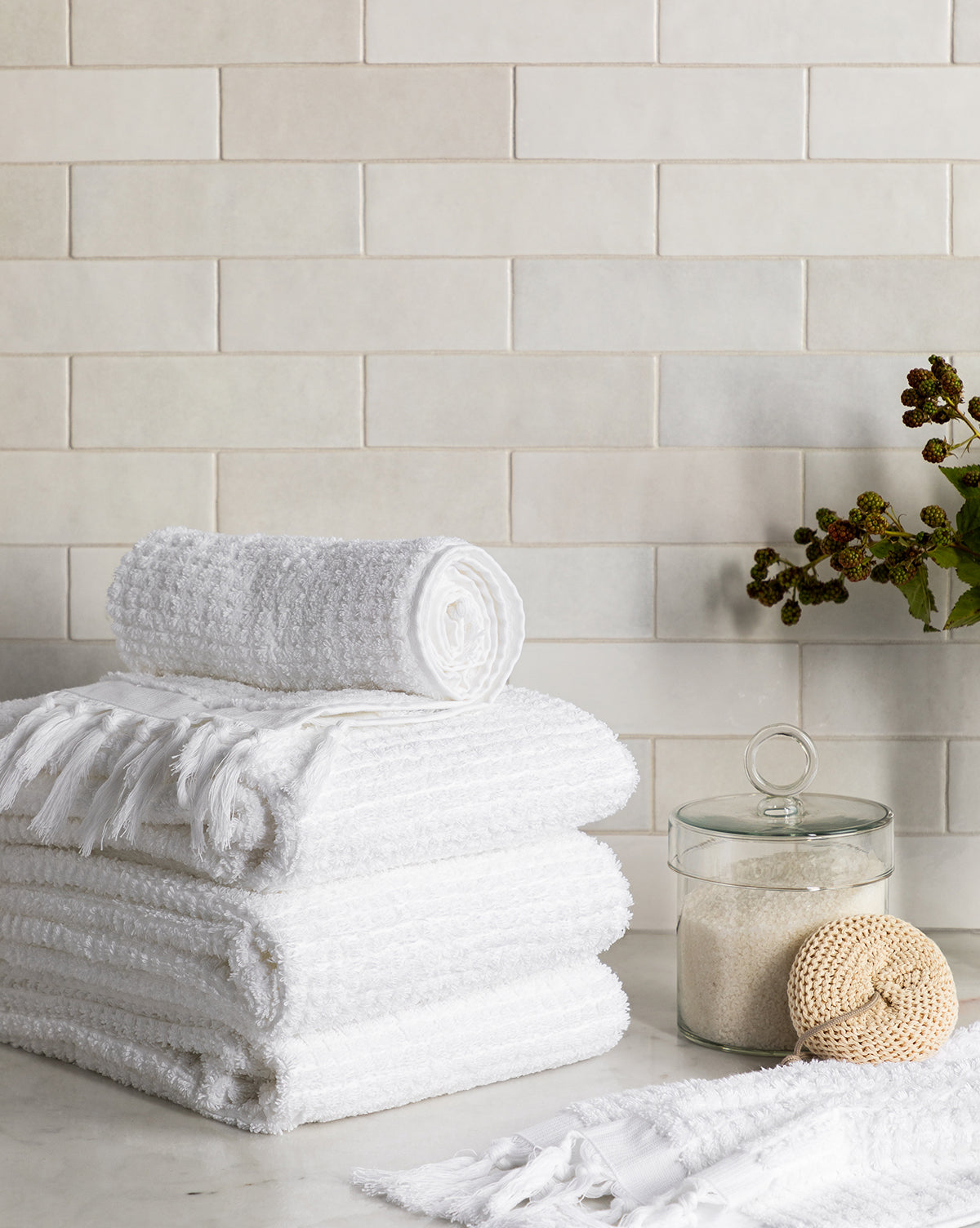 A stack of white towels, a rolled towel, a Glass Lidded Canister with bath powder and round handle, a loofah, and a branch with dark berries rest on a bathroom countertop against light tiled walls.