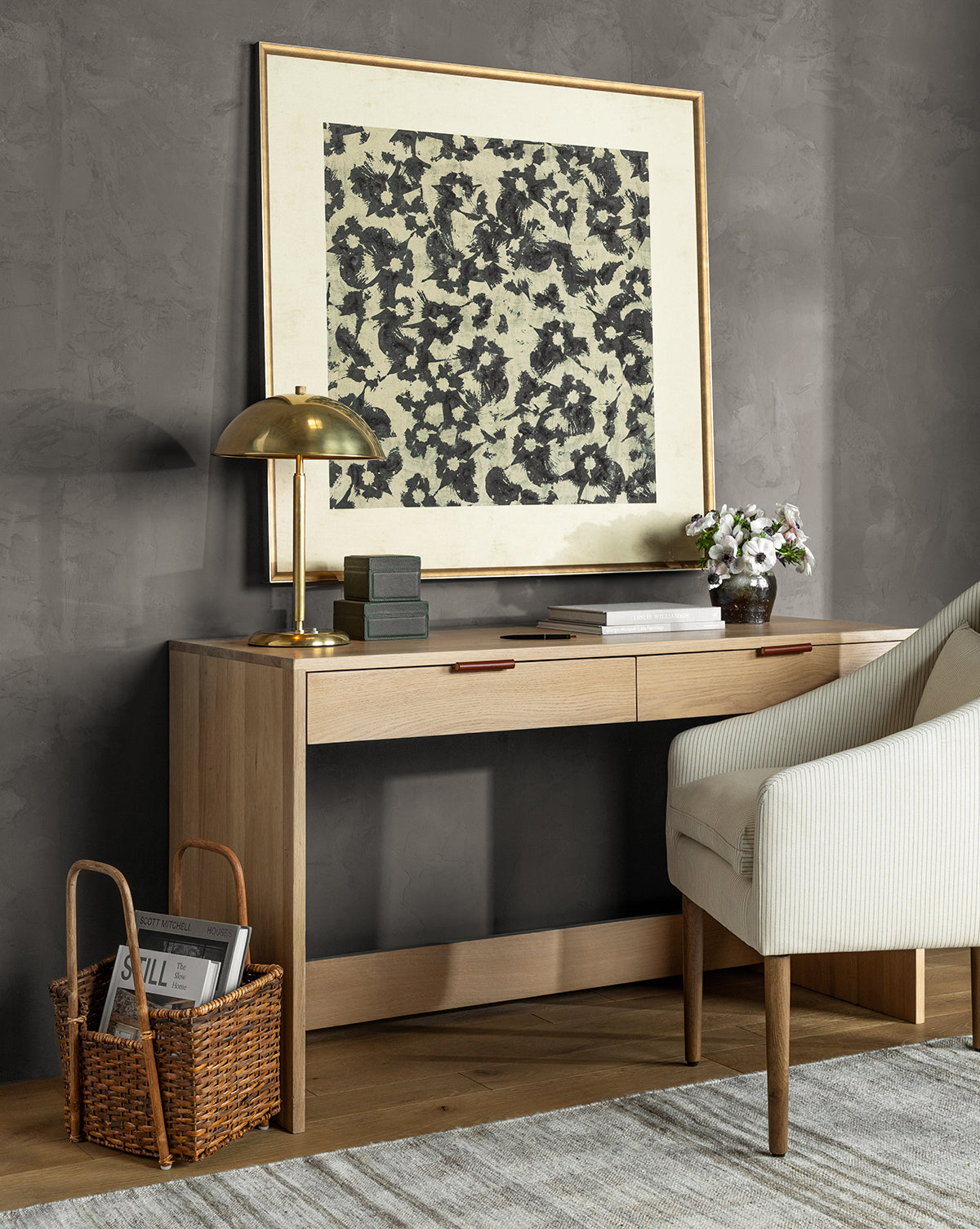 A modern wooden desk with books, a brass lamp, and flowers sits against a gray wall. Above, the Woodblocked Botanical giclee print in a gold frame hangs. Nearby are a striped chair, magazine basket, and light rug.