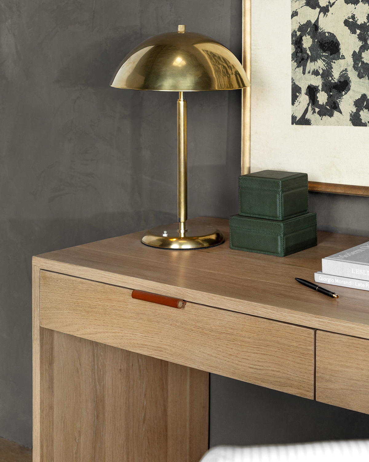 A modern wooden desk features a brass dome-shaped lamp, two stacked Green Leather Boxes, a closed laptop, and framed floral artwork set against a gray wall.