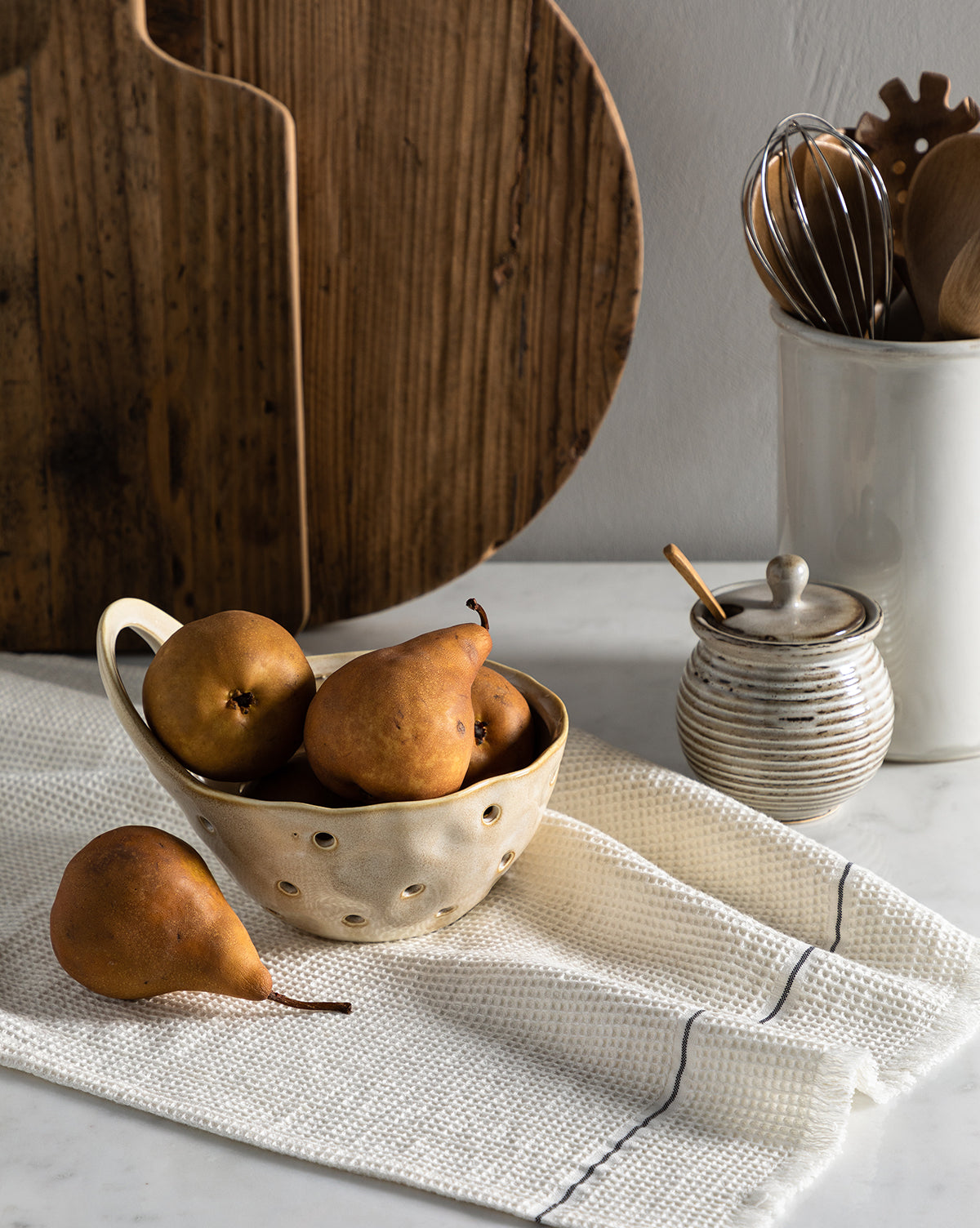 Ripe brown pears in a ceramic colander sit atop the Waffle Hand Towel, with one pear outside. Behind them are a wooden board, a ceramic utensil holder filled with kitchen tools, and a small sugar jar, by McGee & Co.