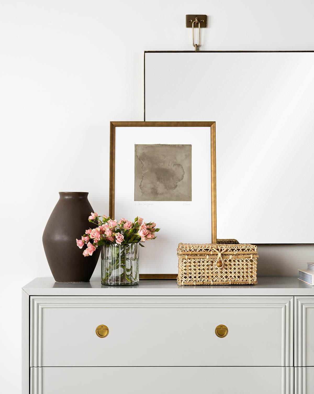 The Olive Study dresser, featuring gold drawer pulls, displays a brown vase, a pink floral glass vase, abstract framed art, books, and a woven box beneath a textured art print and large mirror on a white wall.