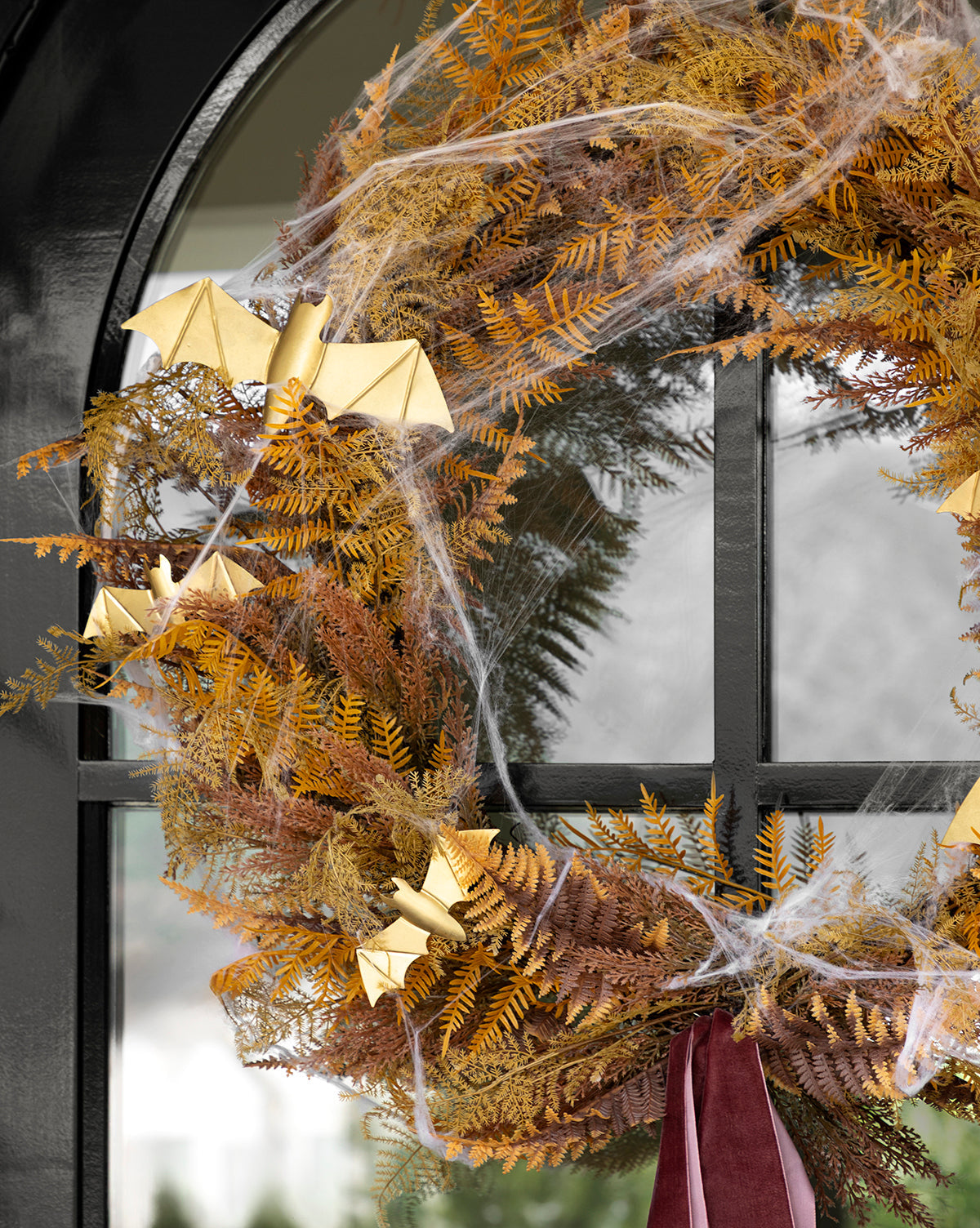 The 30 Faux Golden Fern Wreath hangs on a black door, featuring dried autumn leaves, gold origami bats, artificial cobwebs, and a burgundy ribbon. Ideal for fall entryway decor, by McGee & Co.
