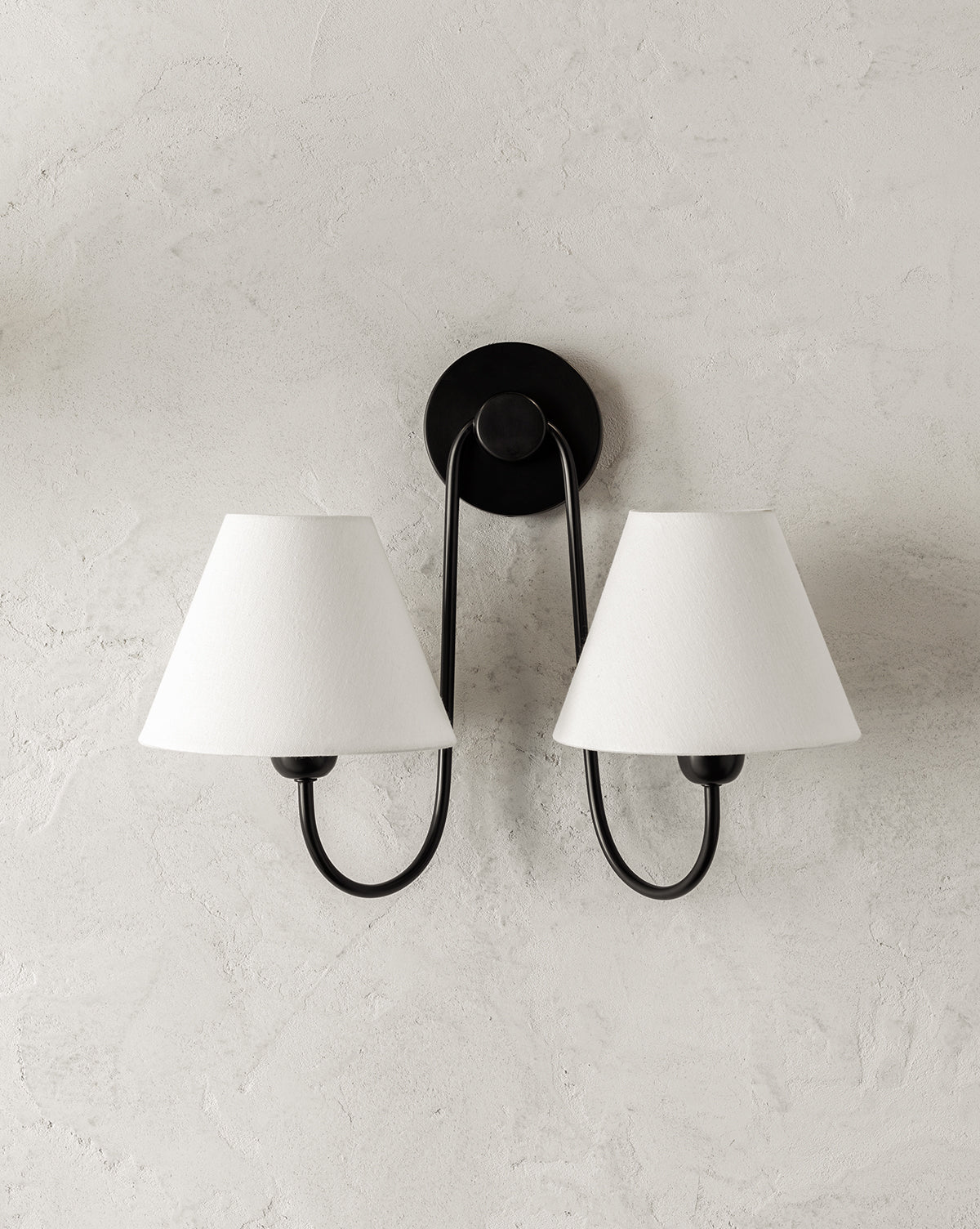 The Wainwright Double Swoop Sconce by Makerspalm features two black metal arms and white linen shades, elegantly mounted on a light textured wall. McGee & Co.