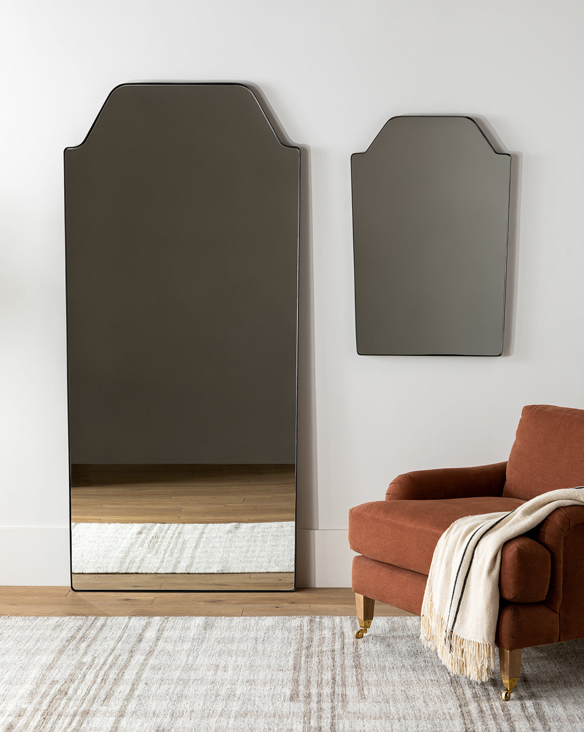 A modern room features the tall matte black Giles Floor Mirror with a geometric top, paired with a matching wall mirror, beside a rust armchair draped in a light throw on a neutral rug over wood flooring, by McGee & Co.