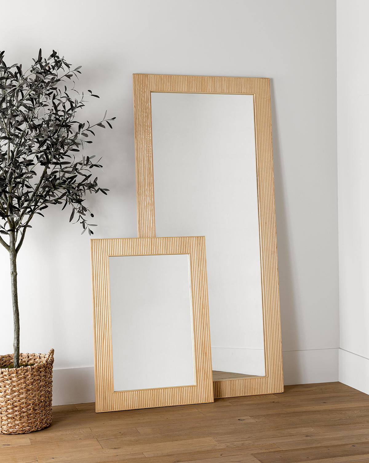 Two Rowan Wall Mirrors with light ribbed ash wood frames lean against a white wall on a wooden floor. A potted plant in a woven basket accents these modern Rowan Wall Mirrors, by McGee & Co.