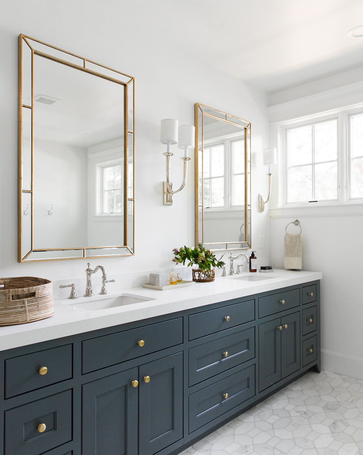 Modern bathroom with double sinks, dark blue cabinets with gold knobs, two Celine Mirrors with burnished gold leaf frames and beveled glass, white countertops, wall sconces, a wicker basket, plants, and a window letting in natural light by McGee & Co.