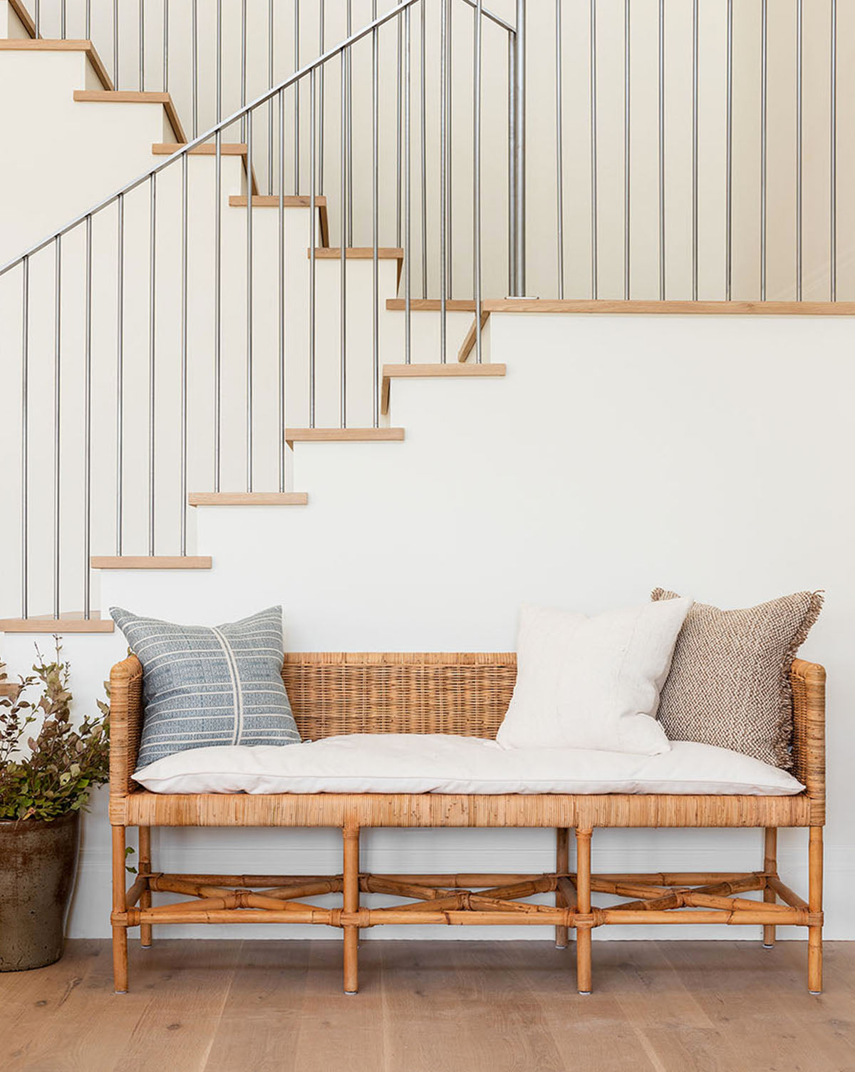 The Odelia Bench with white and patterned cushions sits against a white wall beneath a modern staircase with thin metal railings. A potted plant and a cozy performance fabric pillow add comfort beside the bench.