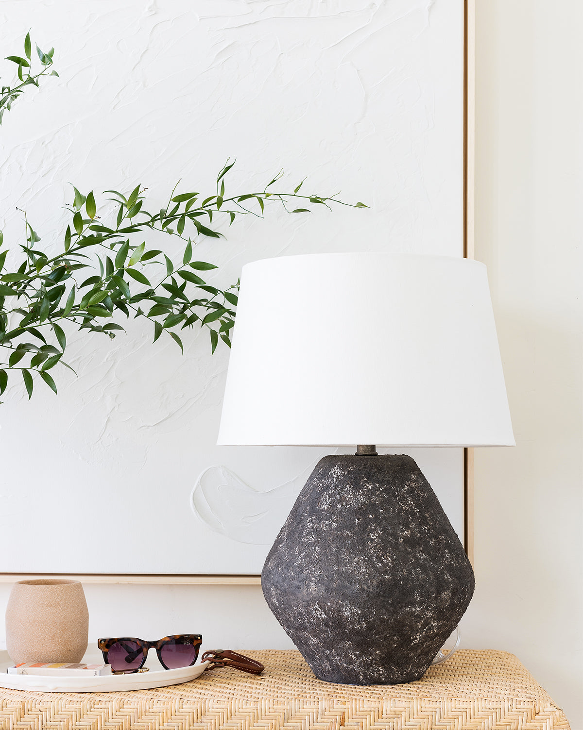 A Rayan Table Lamp with a textured black base and white drum shade sits on a woven table along with a tan cup, sunglasses, and a key, while a leafy green branch is placed before white textured wall art by McGee & Co.