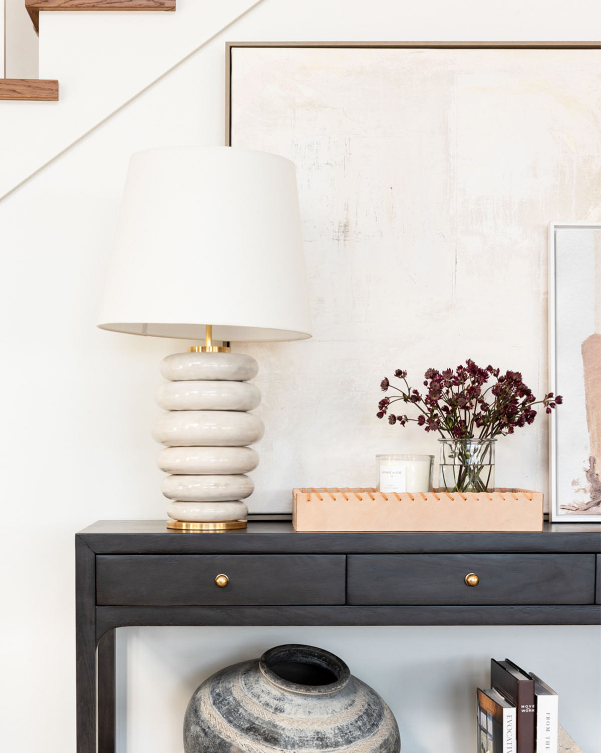 A modern entryway displays the Phoebe Stacked Table Lamp in antiqued white, paired with a vase of dark red flowers, a candle, books, and framed art atop a black console table under the staircase by McGee & Co.