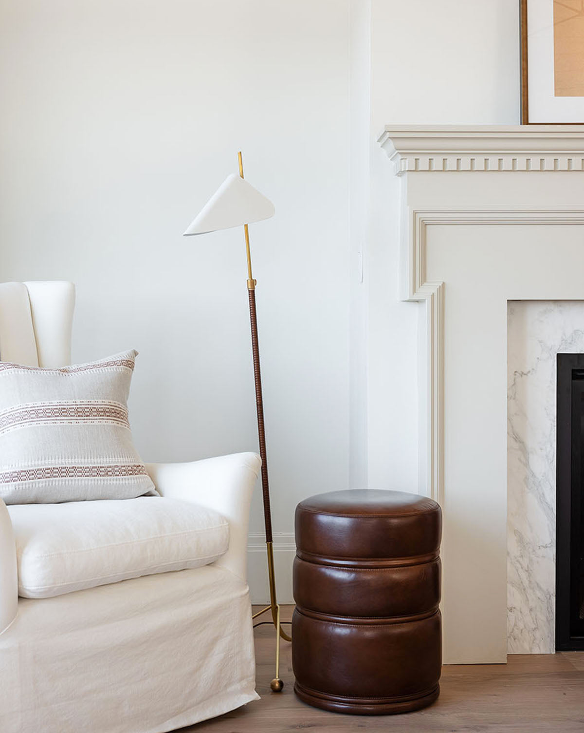 A cozy white armchair with a patterned pillow sits beside the Moresby Floor Lamp, offering stylish lighting, and a round brown leather ottoman is placed near a white fireplace with a marble surround, by McGee & Co.