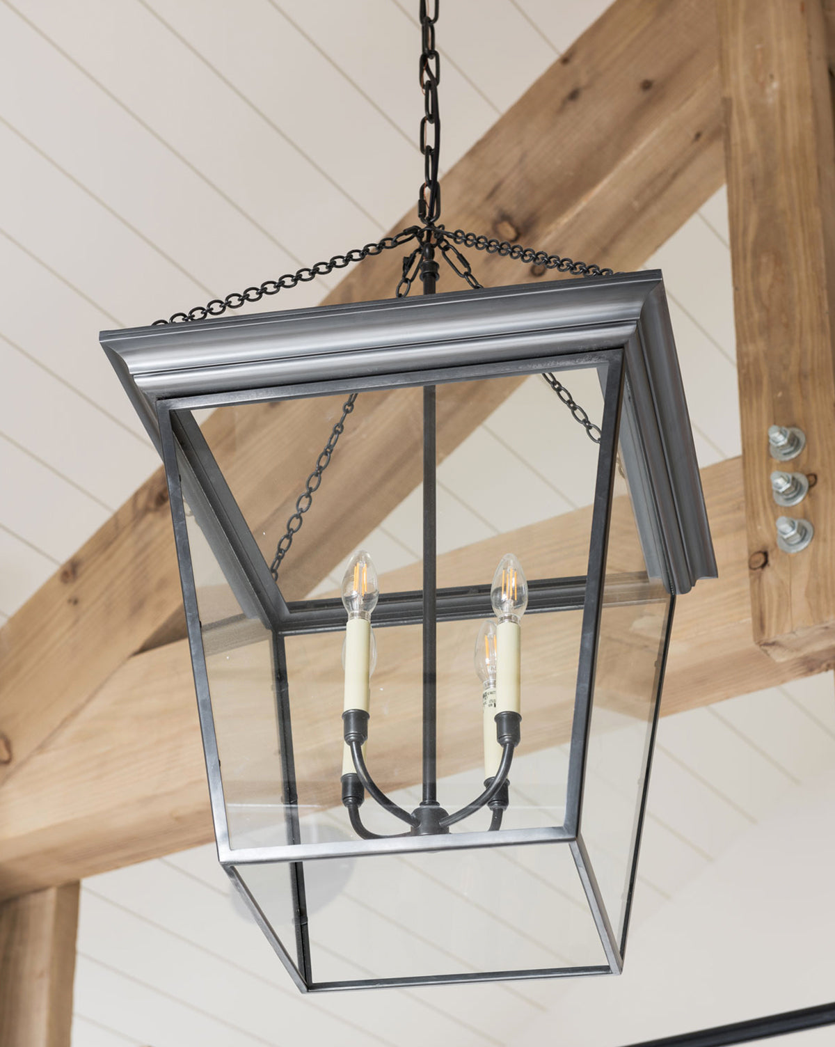 The Cornice Hanging Lantern features a black frame and two candle-style bulbs, suspended by a chain from a wooden ceiling with white panels and exposed beams, merging traditional design with industrial accents, by McGee & Co.