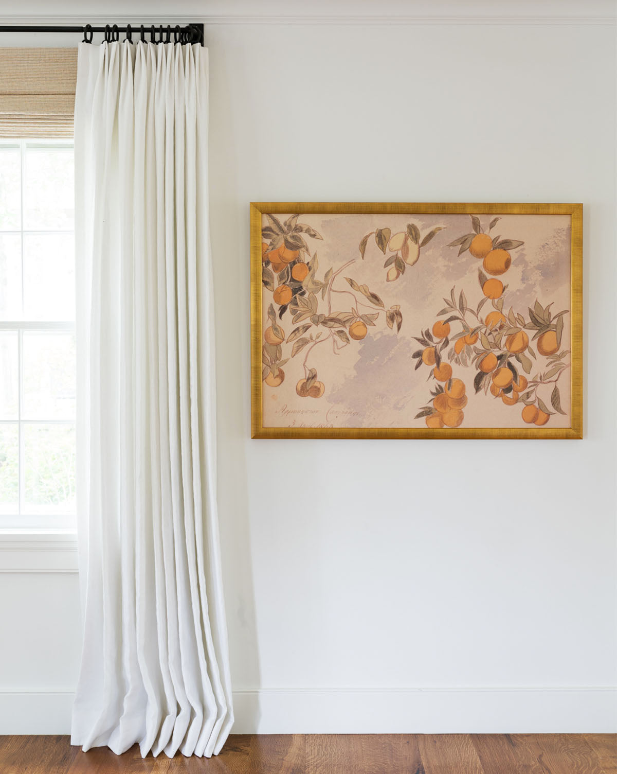 The Lemons & Oranges framed art, featuring vibrant fruit and leaves, hangs on a white wall beside a window with long white curtains. The room has wooden flooring.