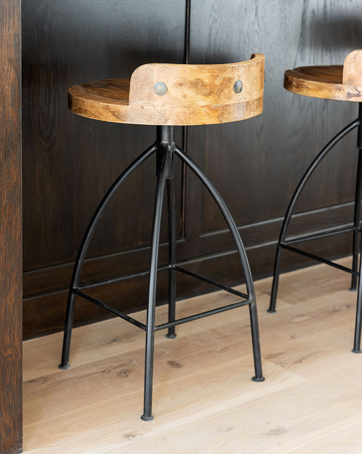 The Johan Counter Stool, featuring a curved wood seat, short backrest, and black metal legs, stands on a light wood floor before a dark wood counter.