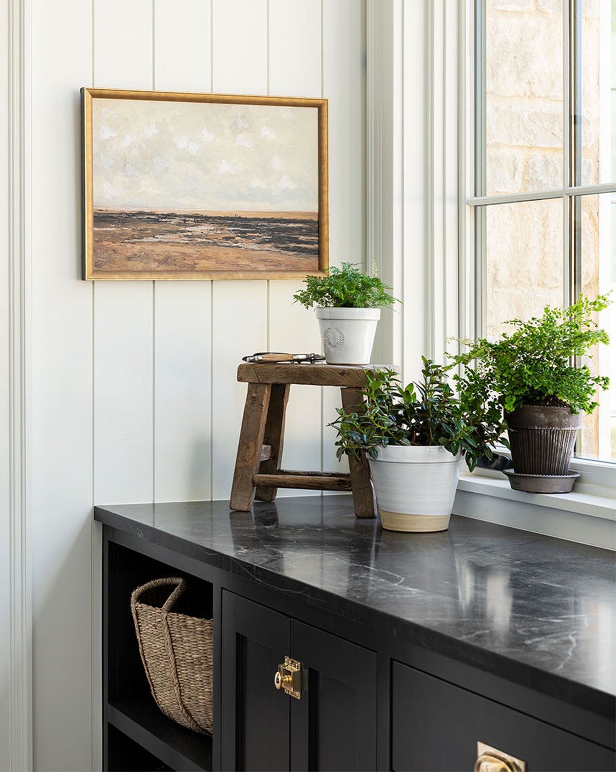 A countertop holds potted plants, a small wooden stool, and a wicker basket. The Villerville Beach framed landscape painting hangs on a white paneled wall near a window with natural light.