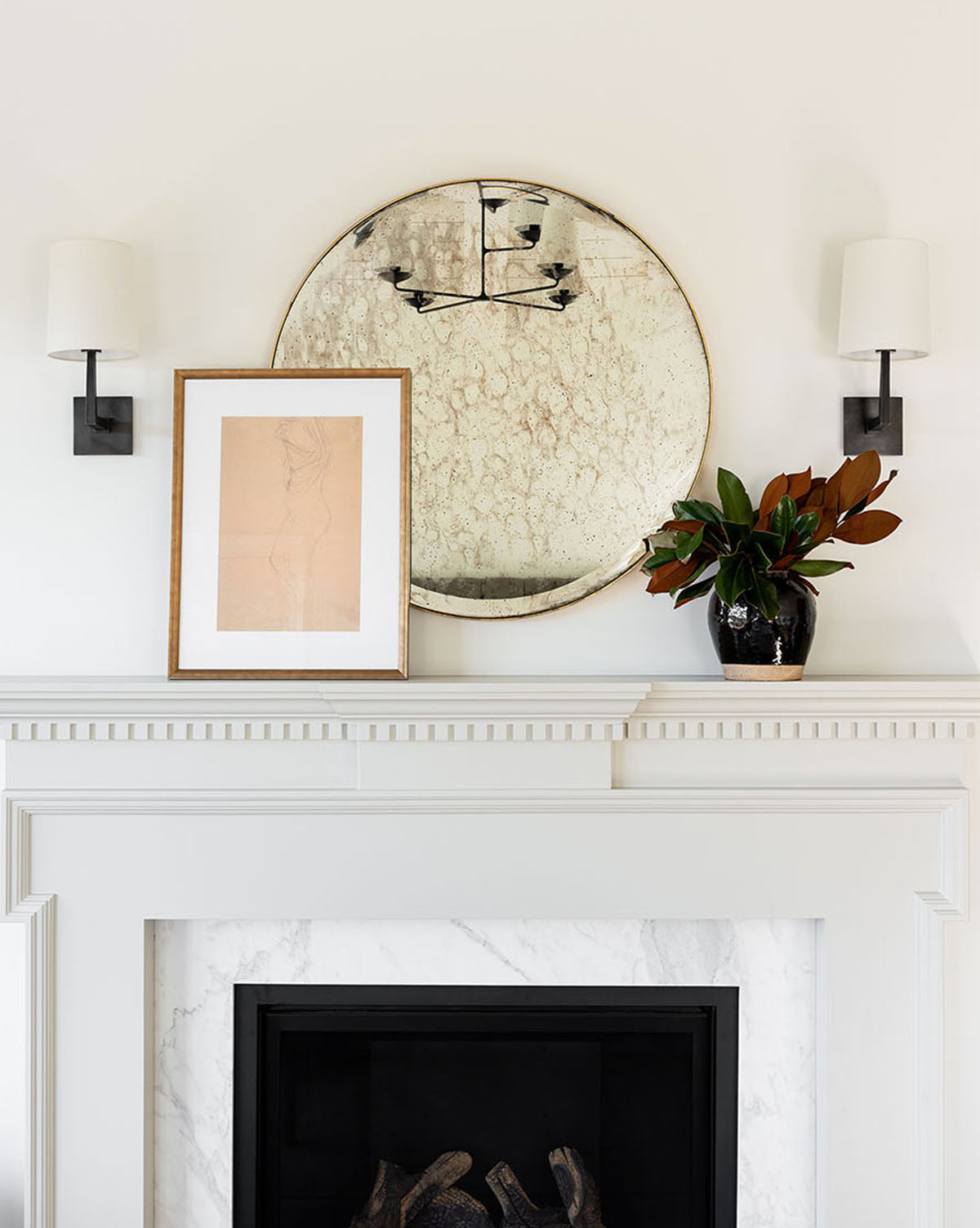 A white fireplace mantel decorated with a round distressed mirror, a simple frame featuring the Sketched Figure artwork, a black vase with leafy branches, and two wall sconces with white shades on either side.