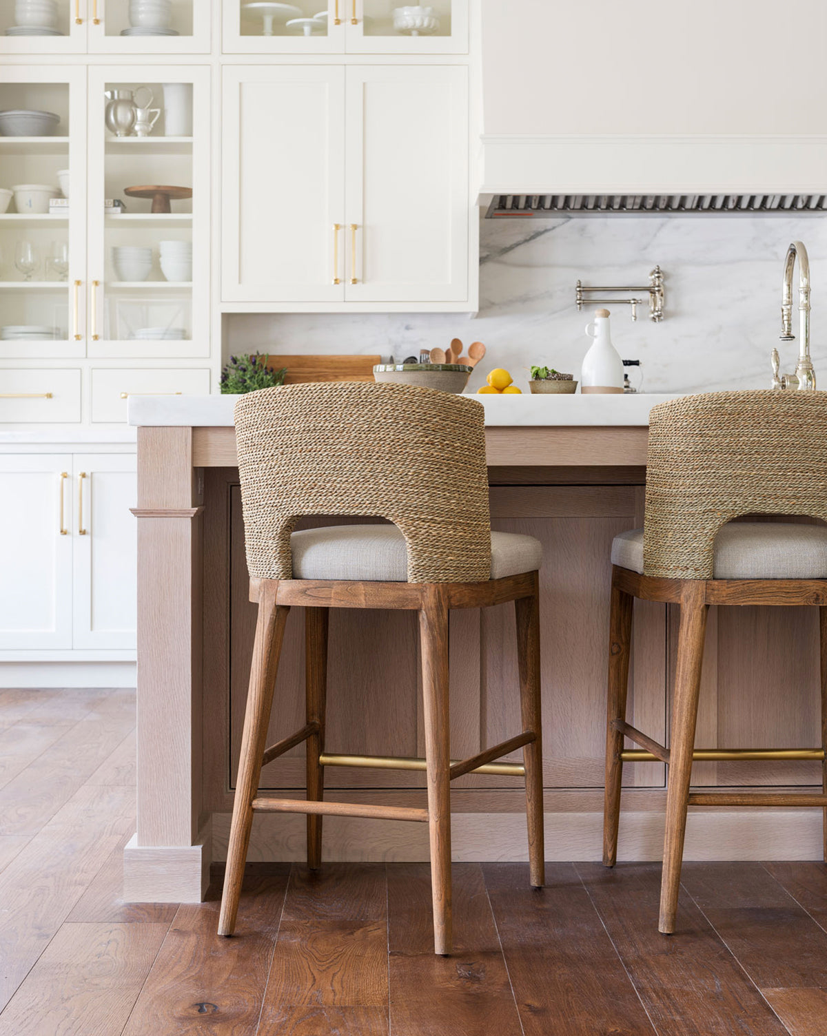 Modern kitchen with white cabinets, glass-front cupboards, and a marble backsplash. Two Ava Seagrass Counter Stools with hand-woven seagrass and teak frames sit at a light wood island on hardwood floors. Essentials and fruit on the countertop.
