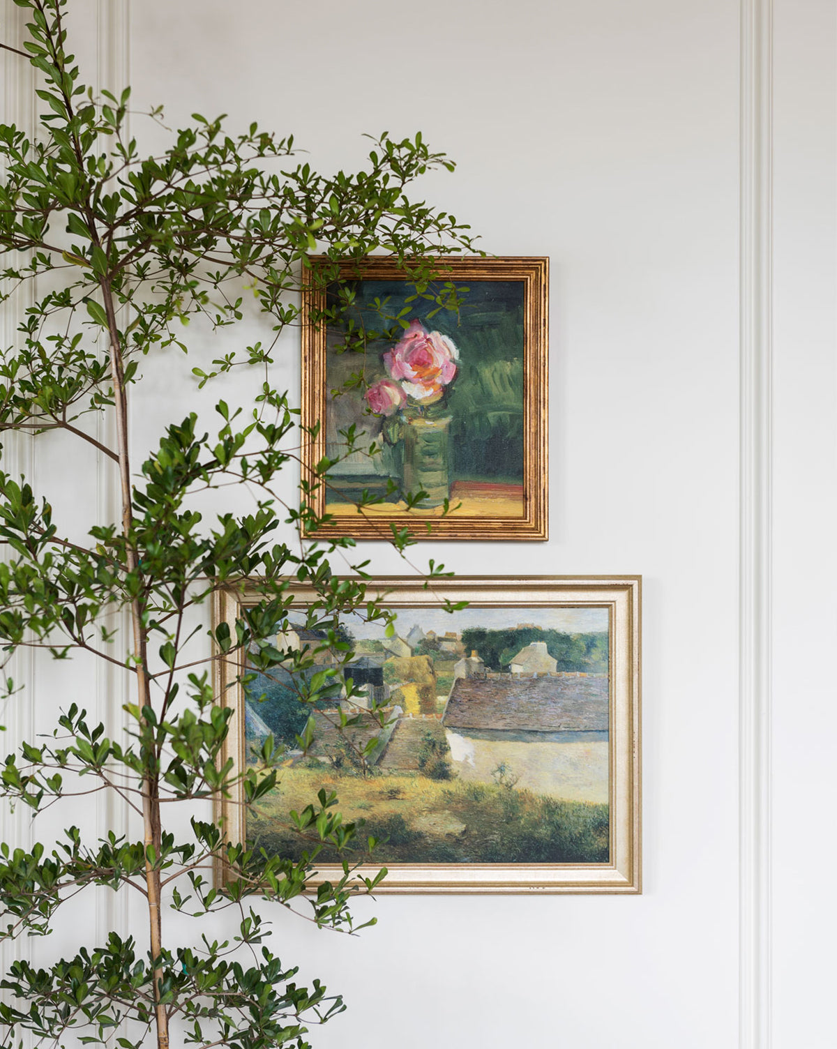 Two framed paintings hang on a white wall: the top shows pink flowers in a vase, while the bottom, Village Impression, features an impressionistic village landscape. A leafy green branch partially obscures the vintage-inspired frames.