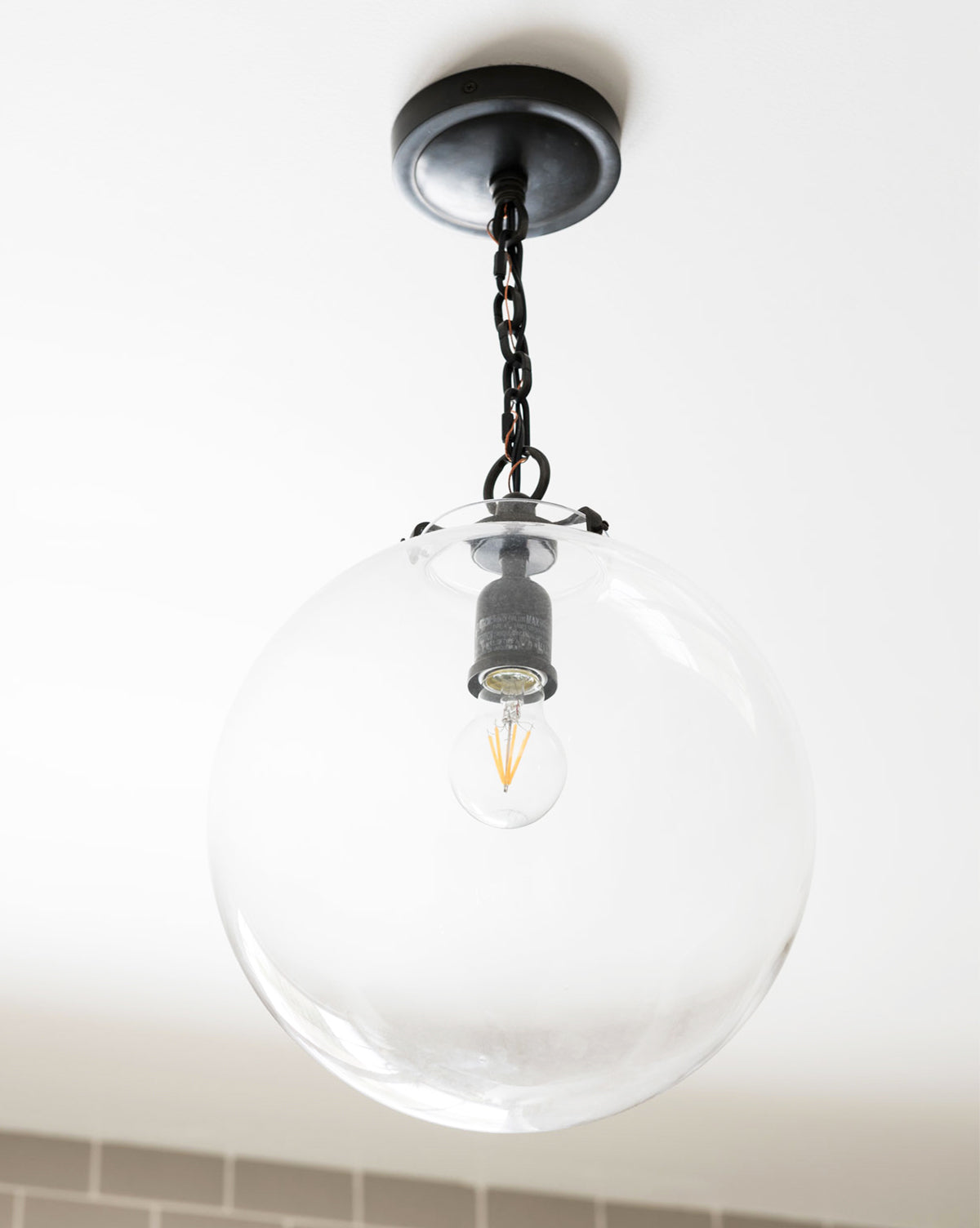 The Katie Globe Pendant by Visual Comfort & Co. Signature features a round glass globe suspended from a short black chain on a white ceiling, highlighting an exposed bulb inside, by McGee & Co.