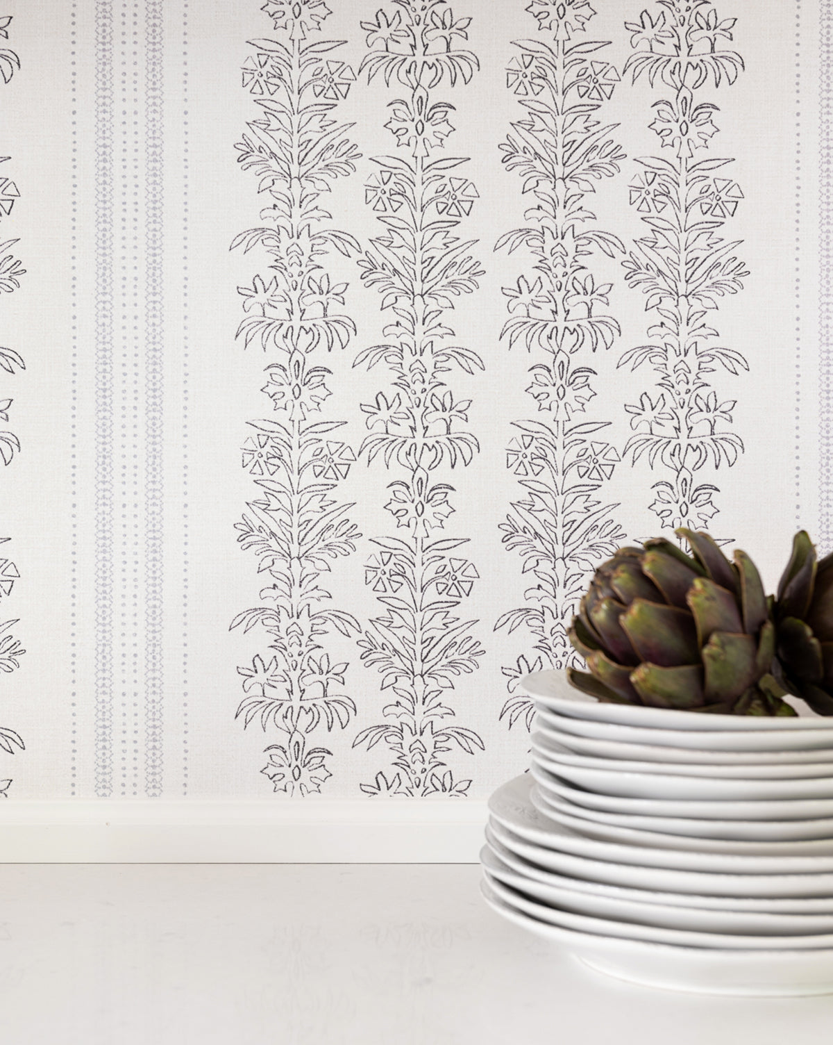 A stack of white plates sits on a white surface, topped with an artichoke, in front of a wall adorned with elegant floral motifs and delicate York Wallcoverings Demi Floral Stripe Wallpaper, McGee & Co.