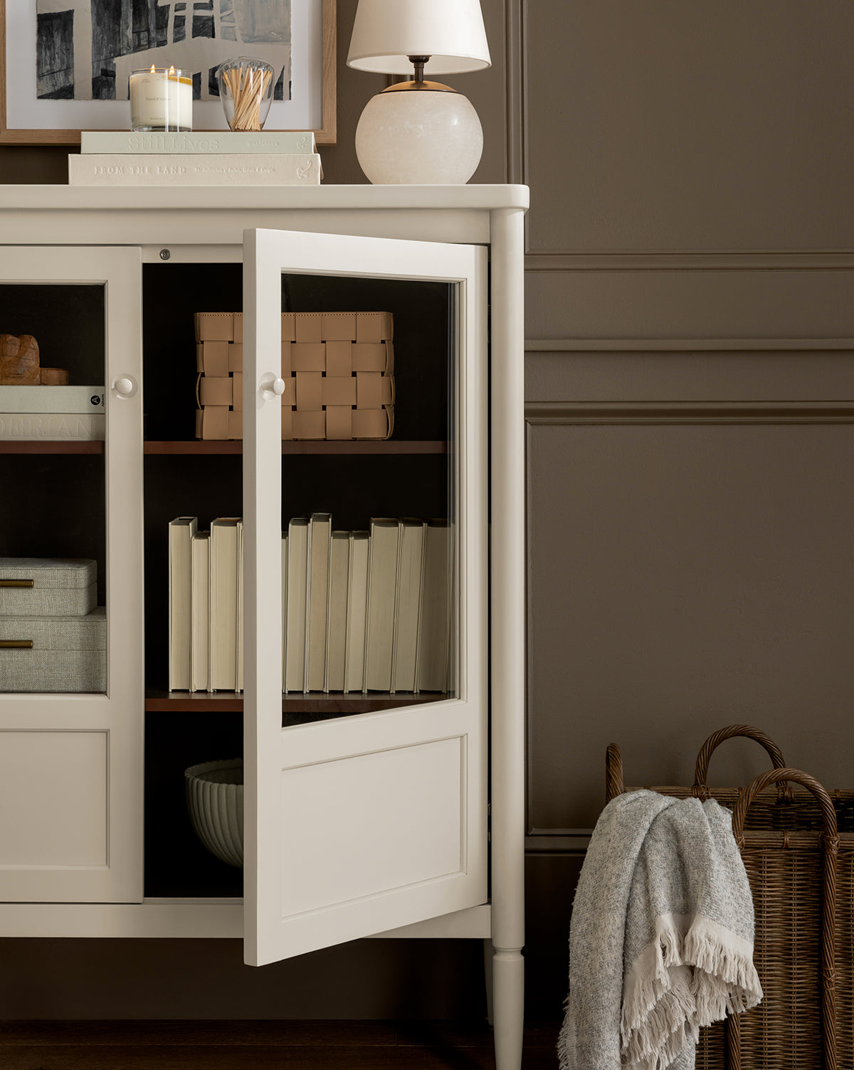 A white cabinet with glass doors stands against a taupe wall in the home office, displaying books and boxes inside. On top are candles, a lamp, and framed art. Beside the cabinet on the floor is the Rupert Leather Bin with a folded blanket.