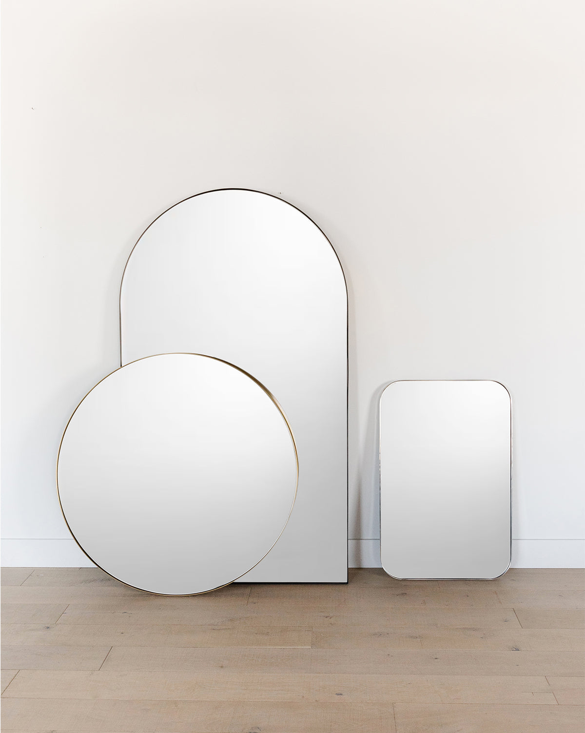 The Jace Inset Rectangle Mirror, featuring shatterproof glass and a frameless design, leans against a plain white wall on a light wooden floor next to two other frameless mirrors—a tall arch and a large circle, by McGee & Co.