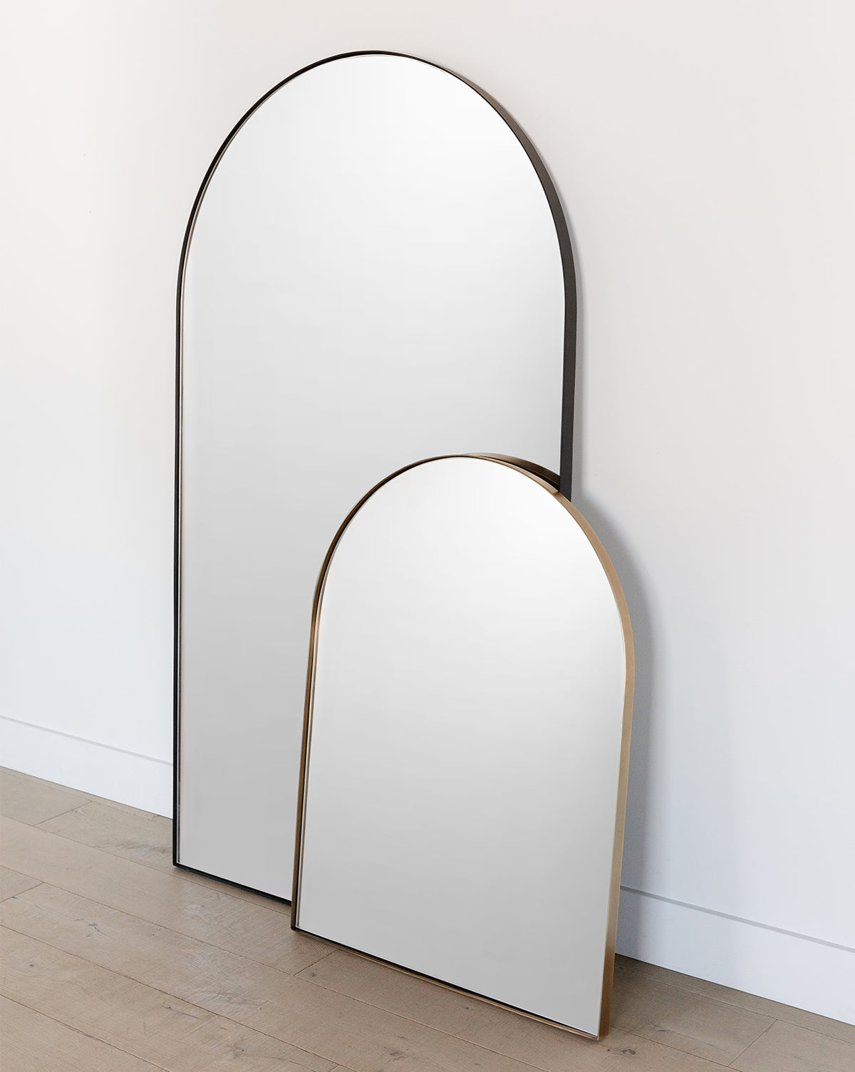 Two Wylie Arched Mirrors with slim metallic frames and shatterproof glass lean against a white wall on a light wooden floor. The taller entryway mirror is partially overlapped by the shorter one, creating a modern layered look, by McGee & Co.