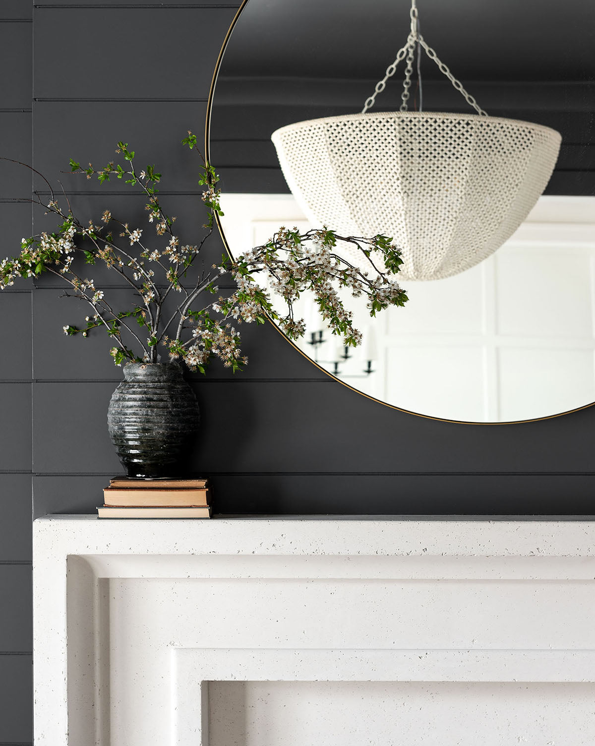 A black vase with flowering branches rests on books atop a white mantel, below the Jace Inset Circle Mirror, which reflects a textured white chandelier against a dark paneled wall, by McGee & Co.