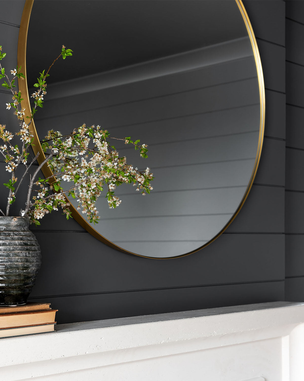 The Jace Inset Circle Mirror with a gold frame hangs on a dark shiplap wall above a white mantel displaying stacked books and a ceramic vase with blooming branches, all reflected in the mirror’s shatterproof glass by McGee & Co.