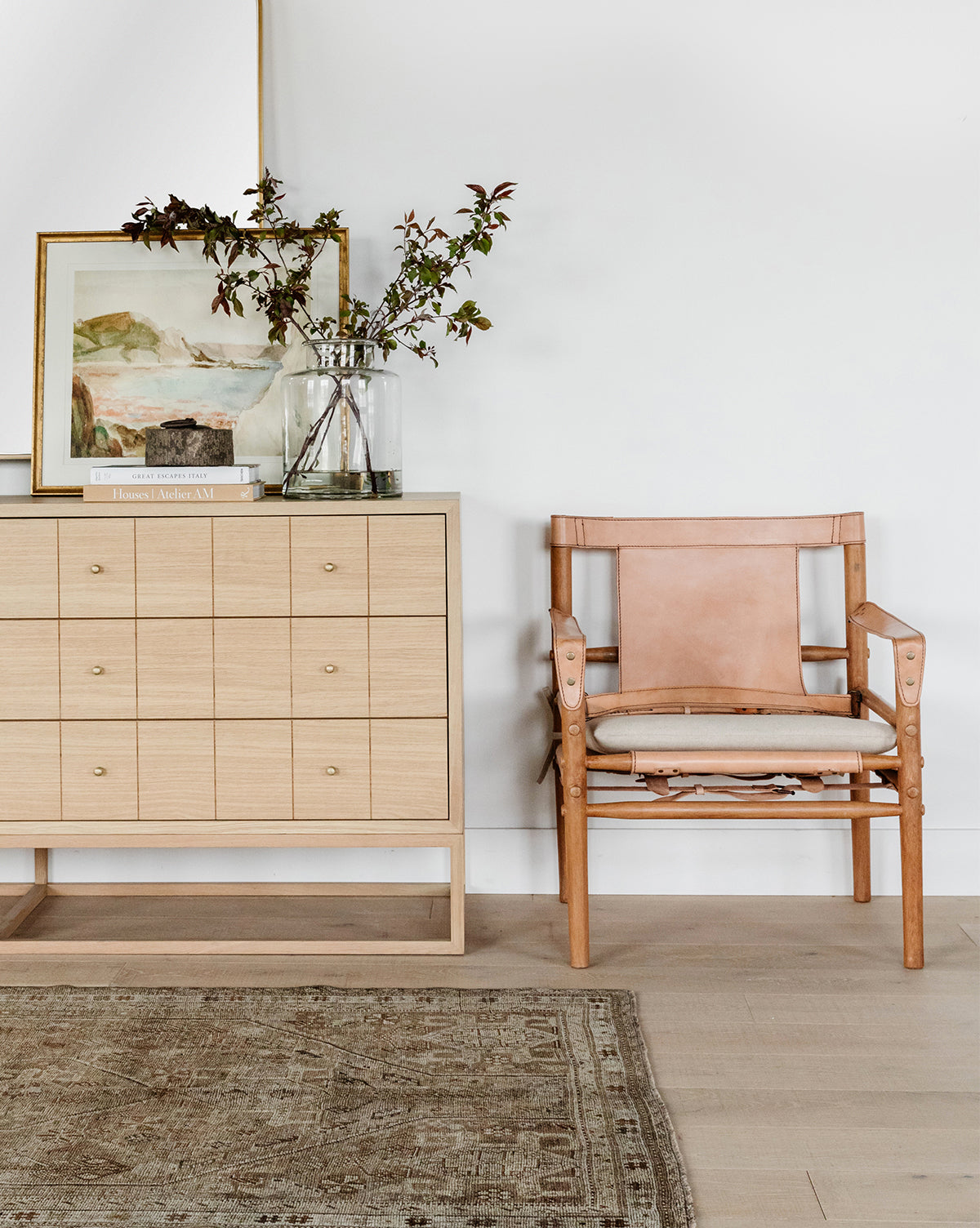 A minimalist room features a light wood sideboard with books, a glass vase of greenery, and the Mountain Reserve decor. Nearby is a tan leather chair on light wood flooring with part of a patterned rug visible.