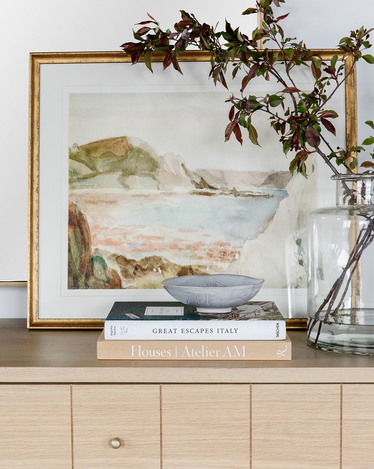 The Mountain Reserve framed wall art, a glass vase with leafy branches, a gray dish, and two books—Great Escapes Italy and Houses | Atelier AM—are displayed on a light wood cabinet.