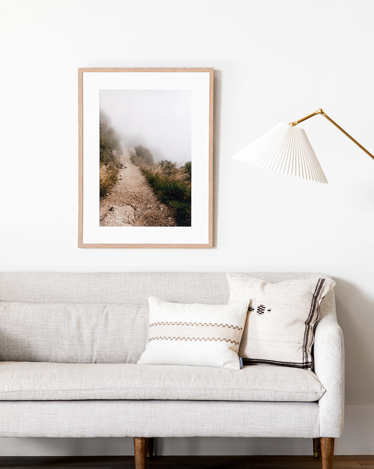 A neutral-toned living room features a light gray sofa with patterned pillows, a gold and white wall lamp, and the framed Foggy Path giclee print hanging on a white wall.