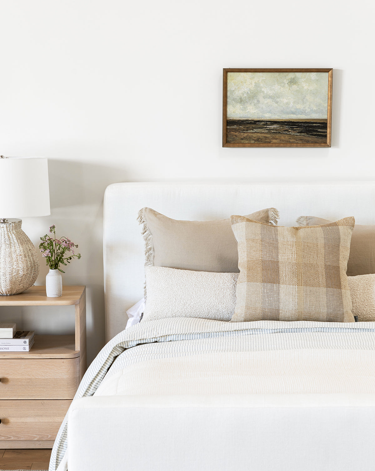 A neatly made bed with neutral bedding and plaid pillows sits beside a wooden nightstand holding a lamp, books, and flowers. Above the bed, the Villerville Beach painting adds charm to the white wall.