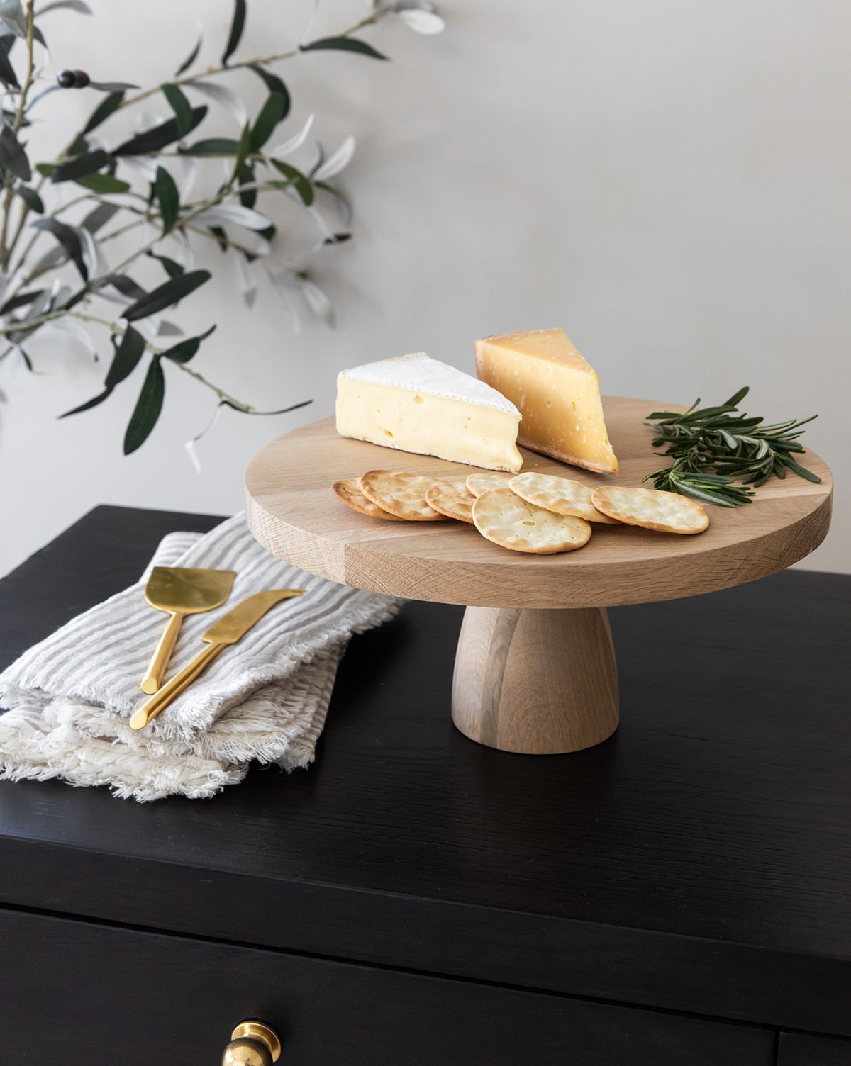 A Wooden Cake Stand displays two cheese wedges, round crackers, and rosemary sprigs. Gold utensils on a folded napkin sit on a dark side table, with leafy branches in the background for stylish home decor by McGee & Co.