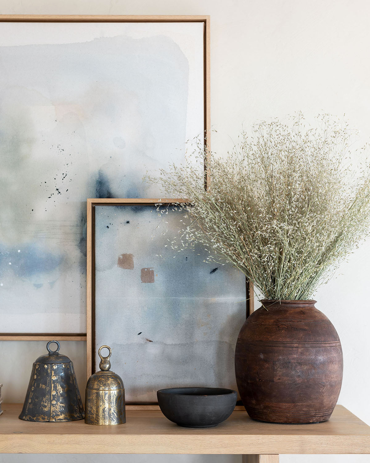 A wooden table showcases two abstract framed artworks, an Aged Wood Vase with dried flowers, a small black bowl, and two metal bells, creating a minimalist scene with built-in styling and soft lighting.