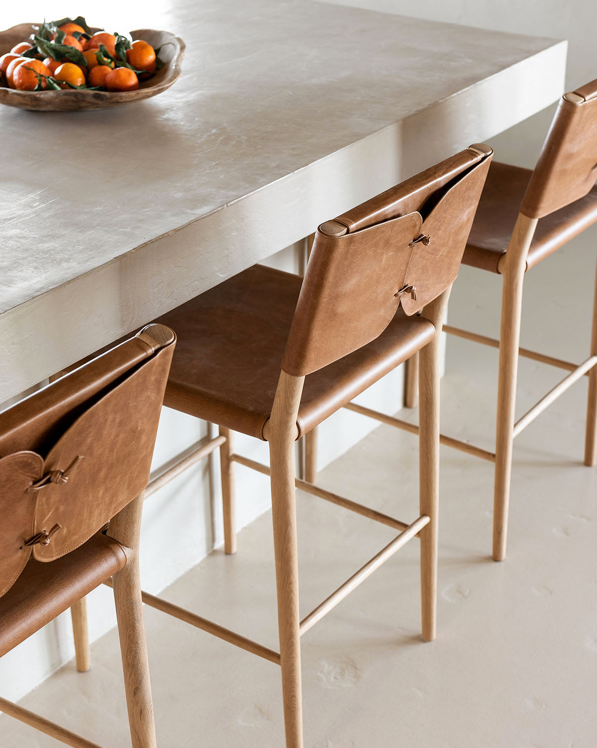 Three Danson Counter Stools with brown leather seats and backs are lined up along a light countertop. A bowl of fresh oranges adds to the modern, minimalist look, by McGee & Co.