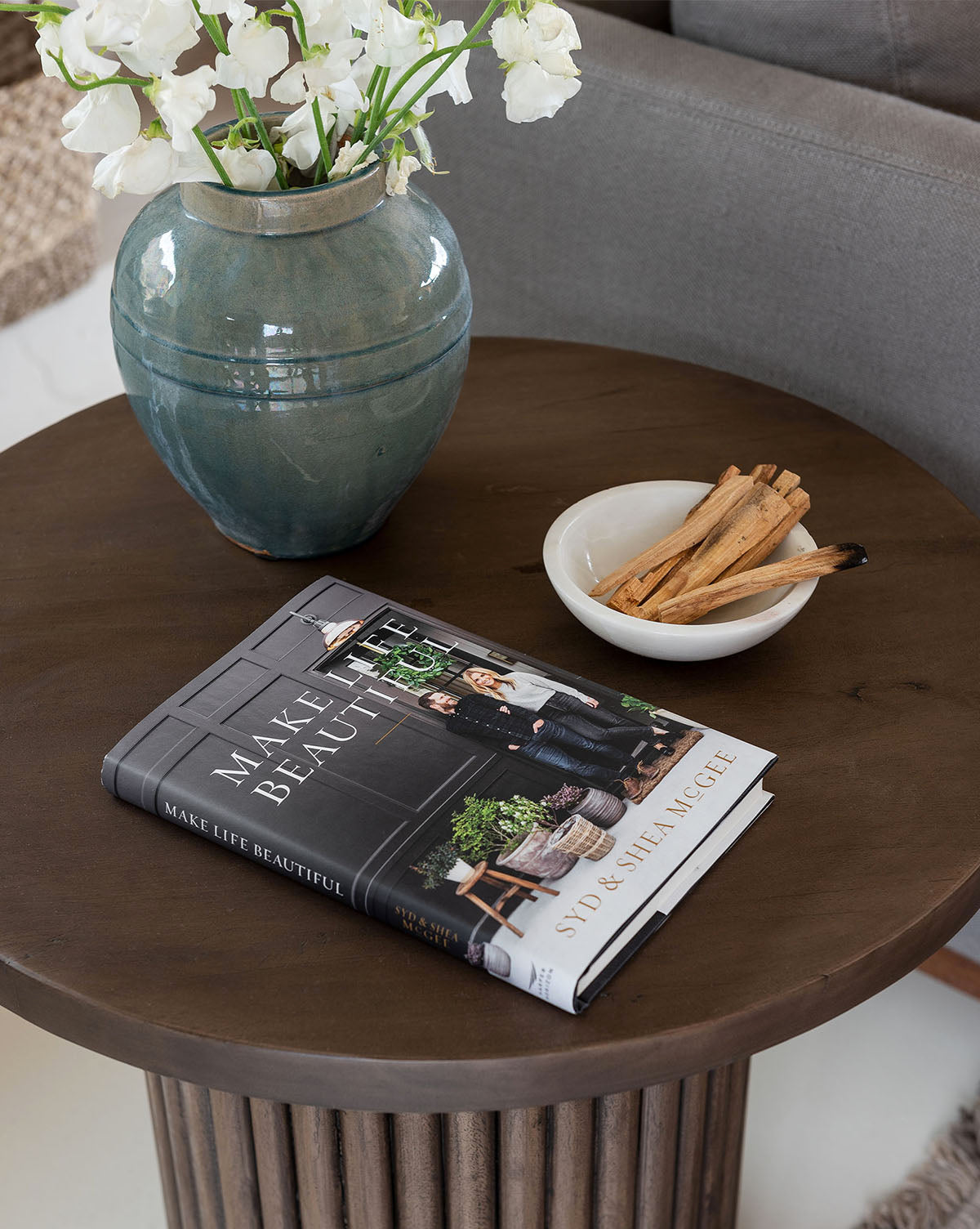 A round wooden side table displays a green vase with white flowers, a set of 3 Marble Nesting Bowls filled with cinnamon sticks, and an open copy of Make Life Beautiful by Syd & Shea McGee for a designer touch.