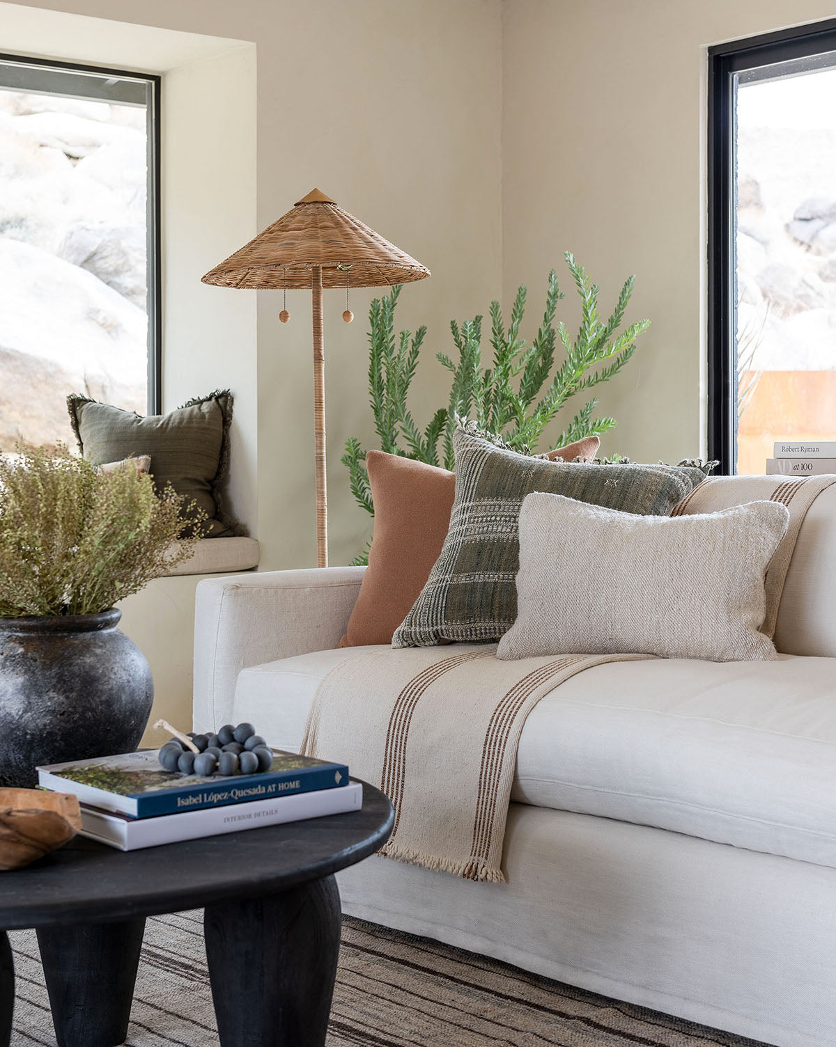 A cozy living room features the Macy Slipcover Sofa (84-96) in white with neutral and green throw pillows, a striped blanket, a black coffee table with books and faux grapes, potted plants, and large windows showcasing a rocky outdoor view by McGee & Co.