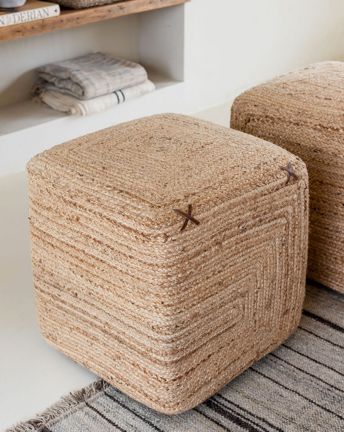 The Kelvin Woven Ottoman, a square hand-woven jute piece, sits on a striped rug beside another pouf. In the background, a wooden shelf displays folded blankets and books for a minimal, cozy feel.