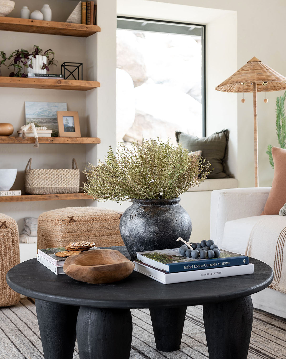 A cozy living room features a black round coffee table, books, a wooden bowl, and the Aged Terracotta Jar with greenery. Woven stools, textured decor on shelves, a white armchair, and a window fill the space with natural light.