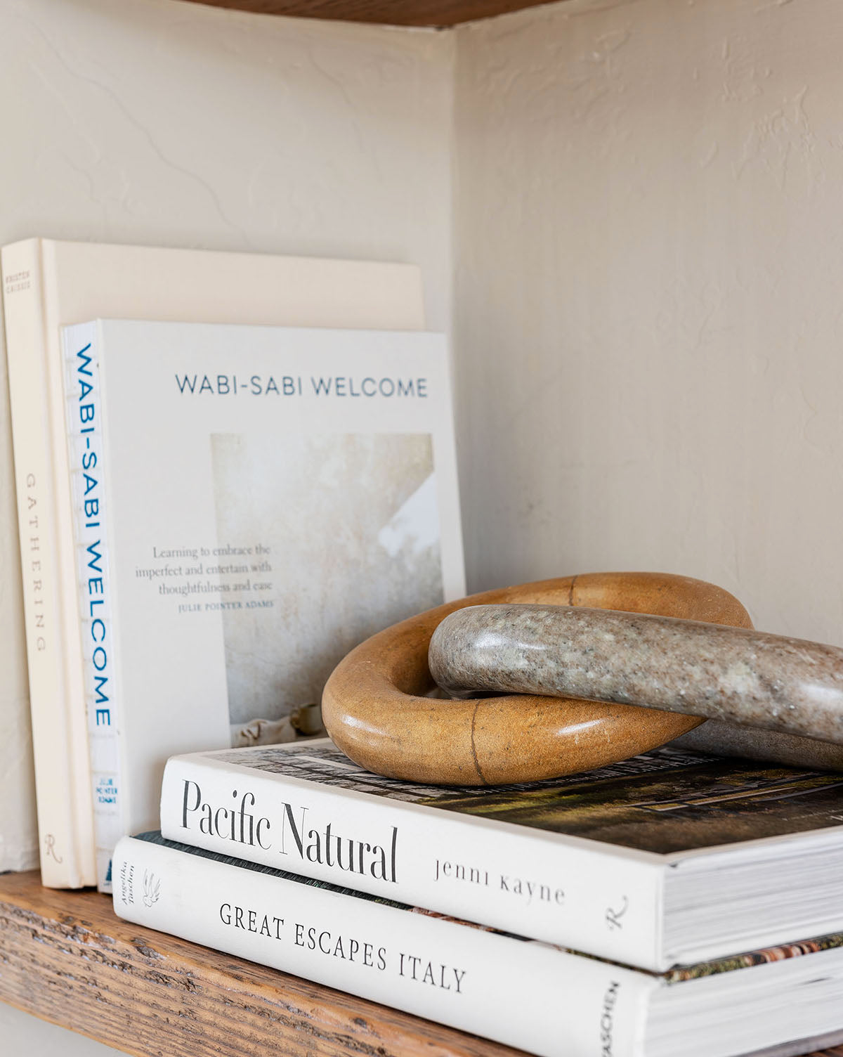 A compact stack of books on a wooden shelf, including Great Escapes Italy—your essential guide to stunning hotels—rests beneath two smooth, stone rings.