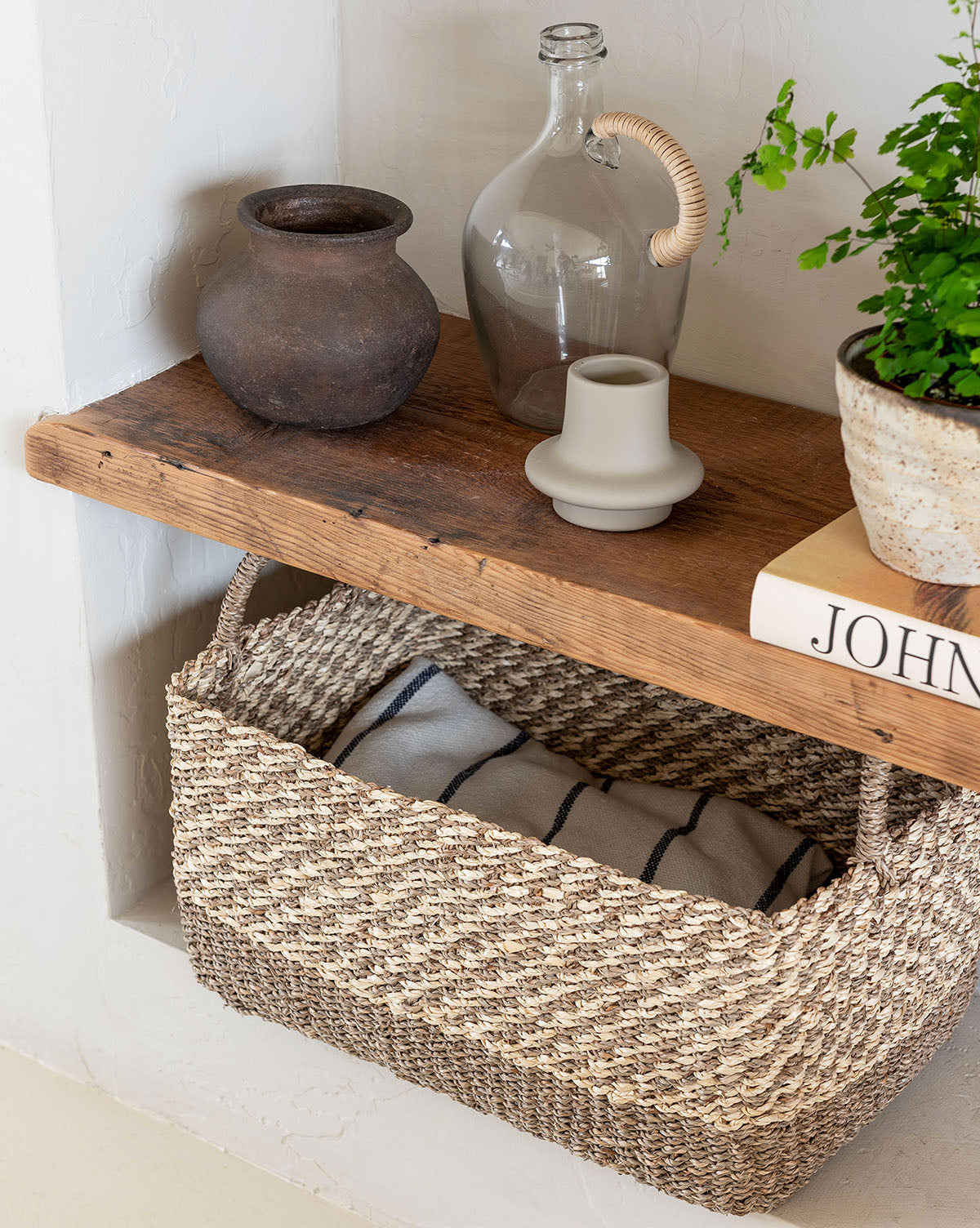 A rustic wooden shelf displays a clay pot, clear glass bottle, beige candle holder, potted plant, and book; beneath the shelf sits the Cordova Basket holding a striped blanket.