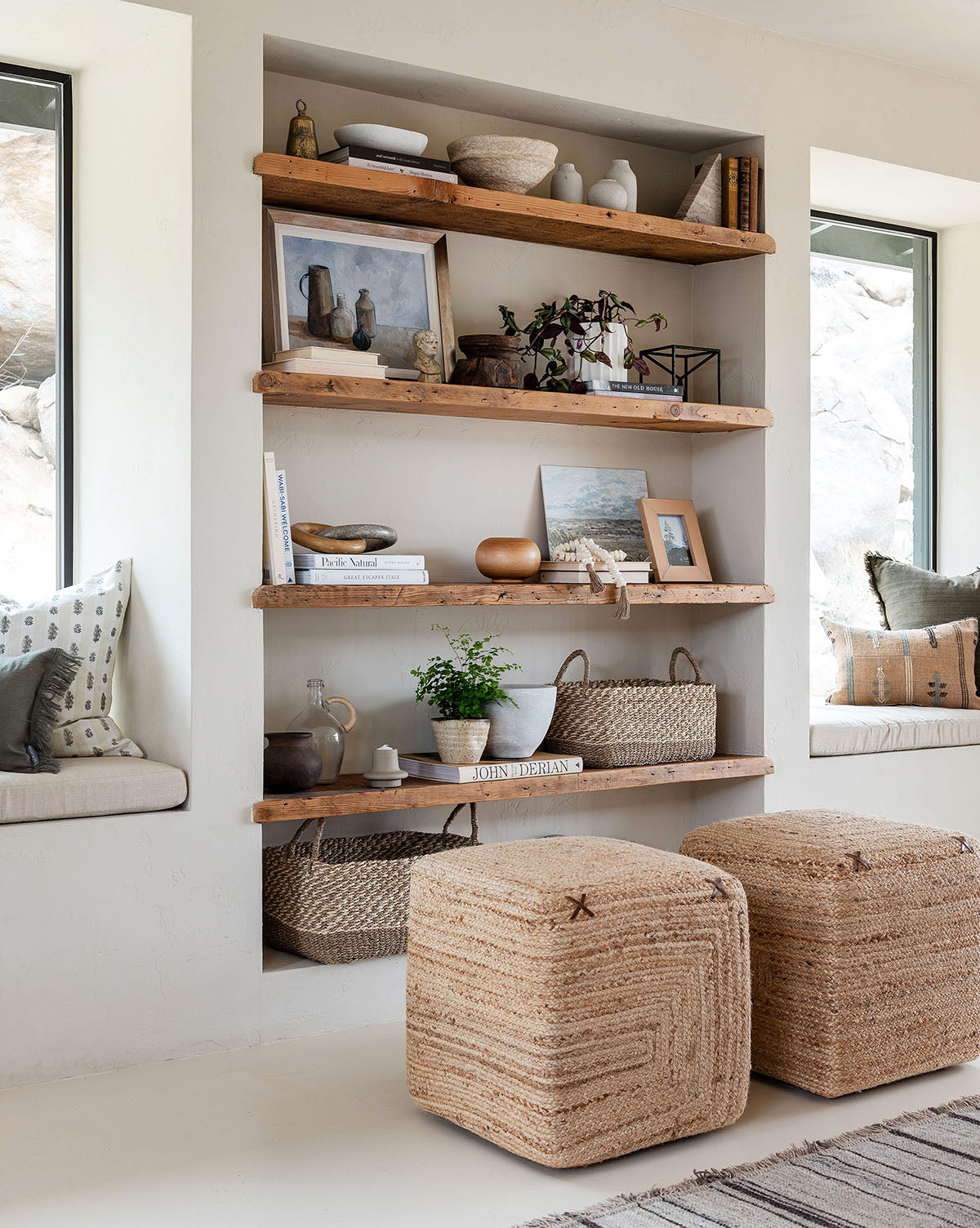 Two Kelvin Woven Ottomans sit in front of built-in wall shelves with rustic wood, which display decor, books, and plants. Cozy window seats with cushions and pillows flank the ottomans as natural light fills the space.