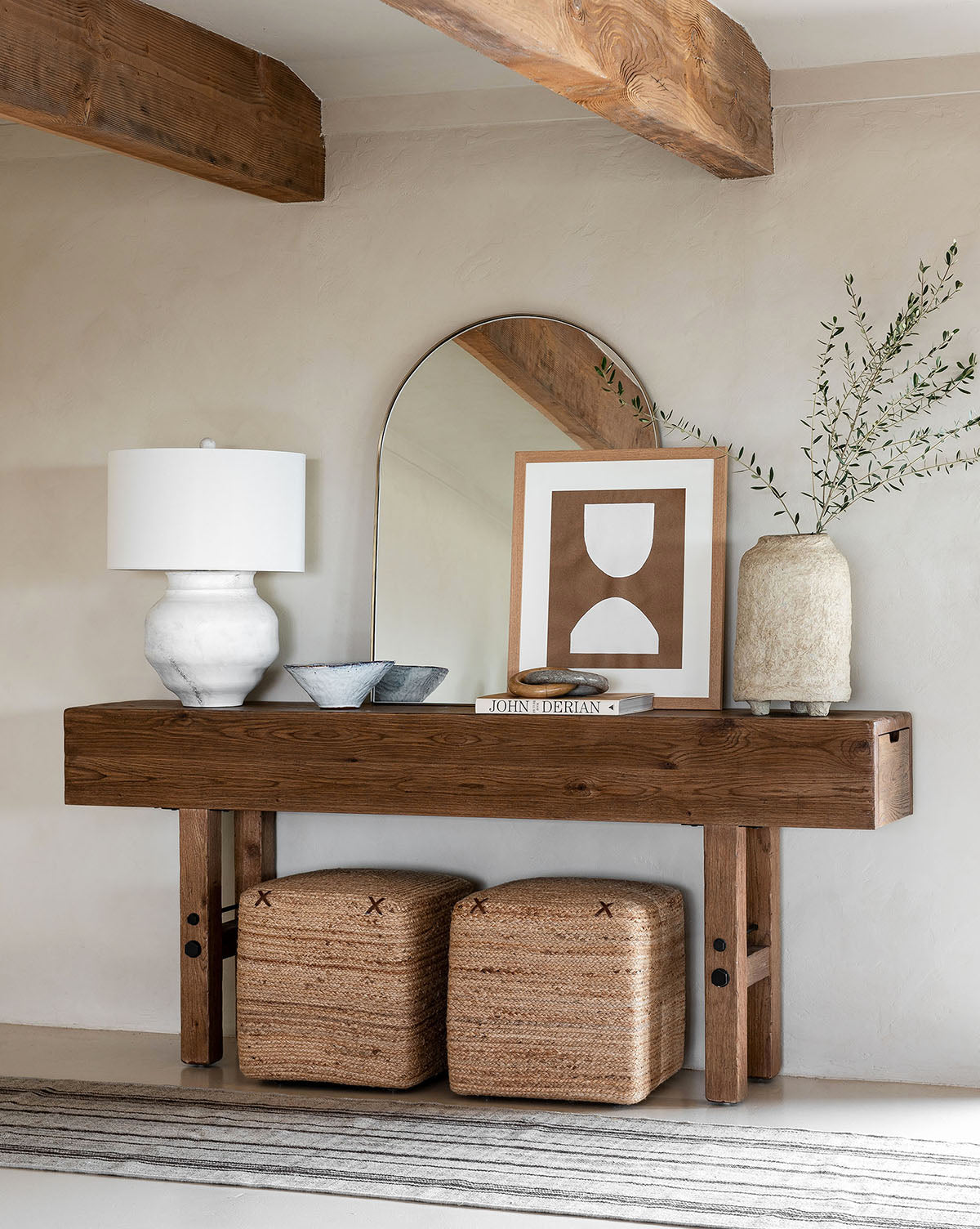 A wooden console table displays a white lamp, bowls, books, framed art, a tall vase with greenery, and an arched mirror. Two Kelvin Woven Ottomans are tucked underneath the table.