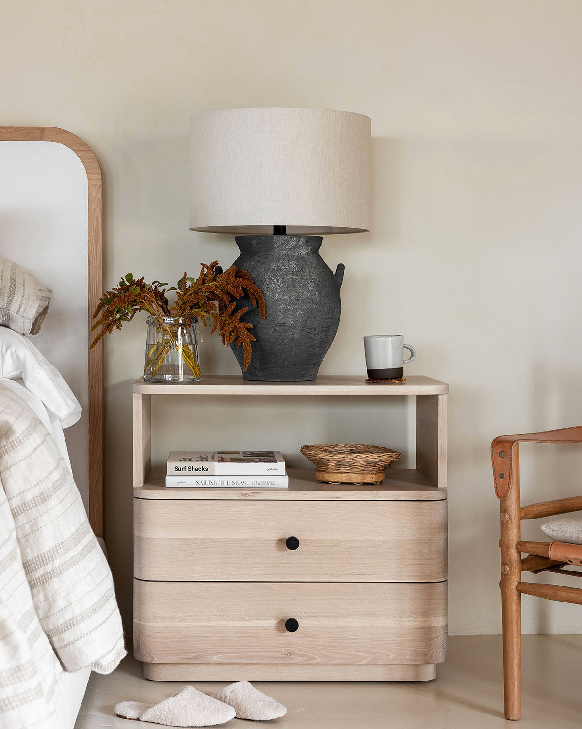 A light wood nightstand next to a bed displays the Anza Table Lamp in black ash glaze, a glass vase with brown foliage, a mug, a basket, and books. Nearby stands a wooden chair draped with a folded blanket by McGee & Co.