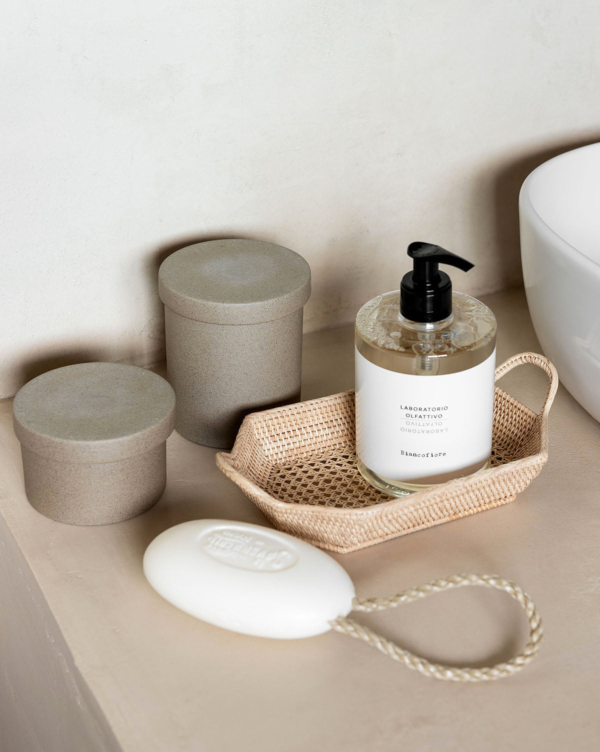 A bottle of Laboratorio Olfattivo Liquid Soap, two beige canisters in a basket, and a white oval botanical soap rest on a neutral countertop next to a white sink.