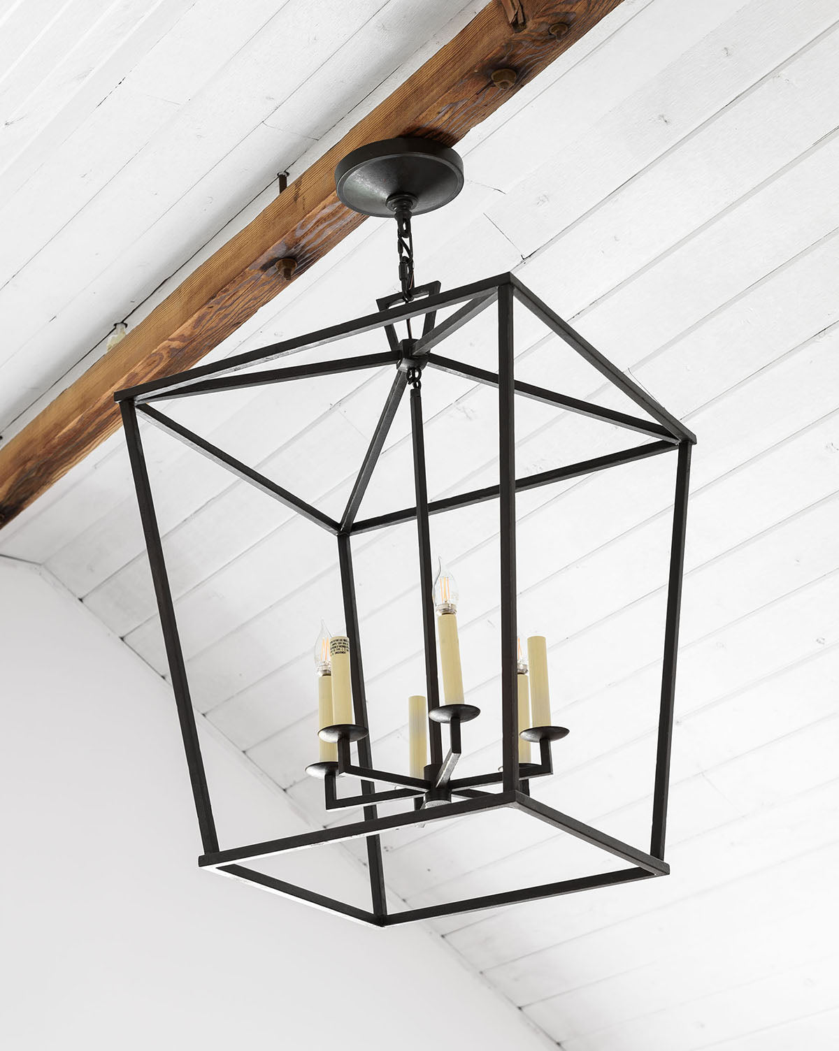 A Darlana Lantern, a black metal contemporary pendant with four candle-style lights, hangs from a white wooden ceiling with exposed dark wood beam, bringing modern style and illumination to the space by McGee & Co.